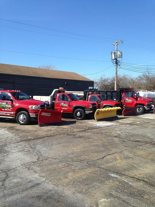 Red snowplow trucks parked in a lot on a sunny day, with a building and utility pole behind them.