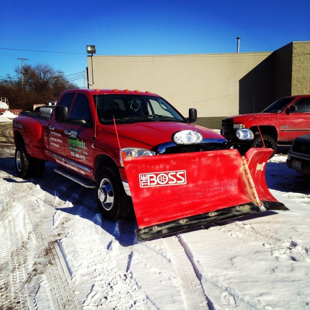 Red pickup truck with a snow plow parked in a snowy lot, with another truck nearby.