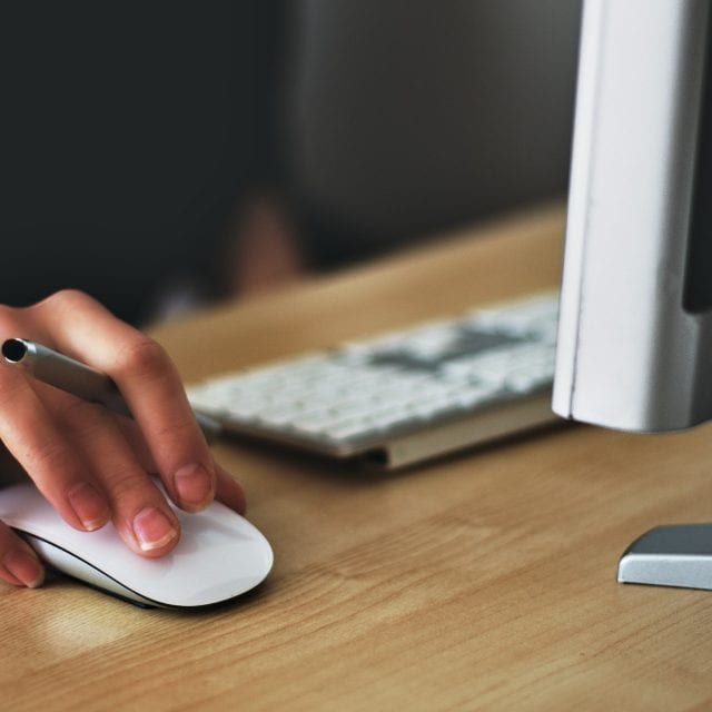 Hand using a mouse beside a keyboard at a desk, with a computer monitor nearby