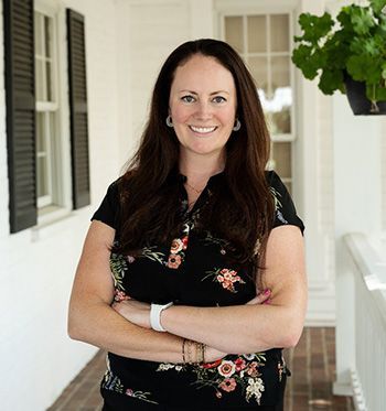 A woman is standing on a porch with her arms crossed and smiling.