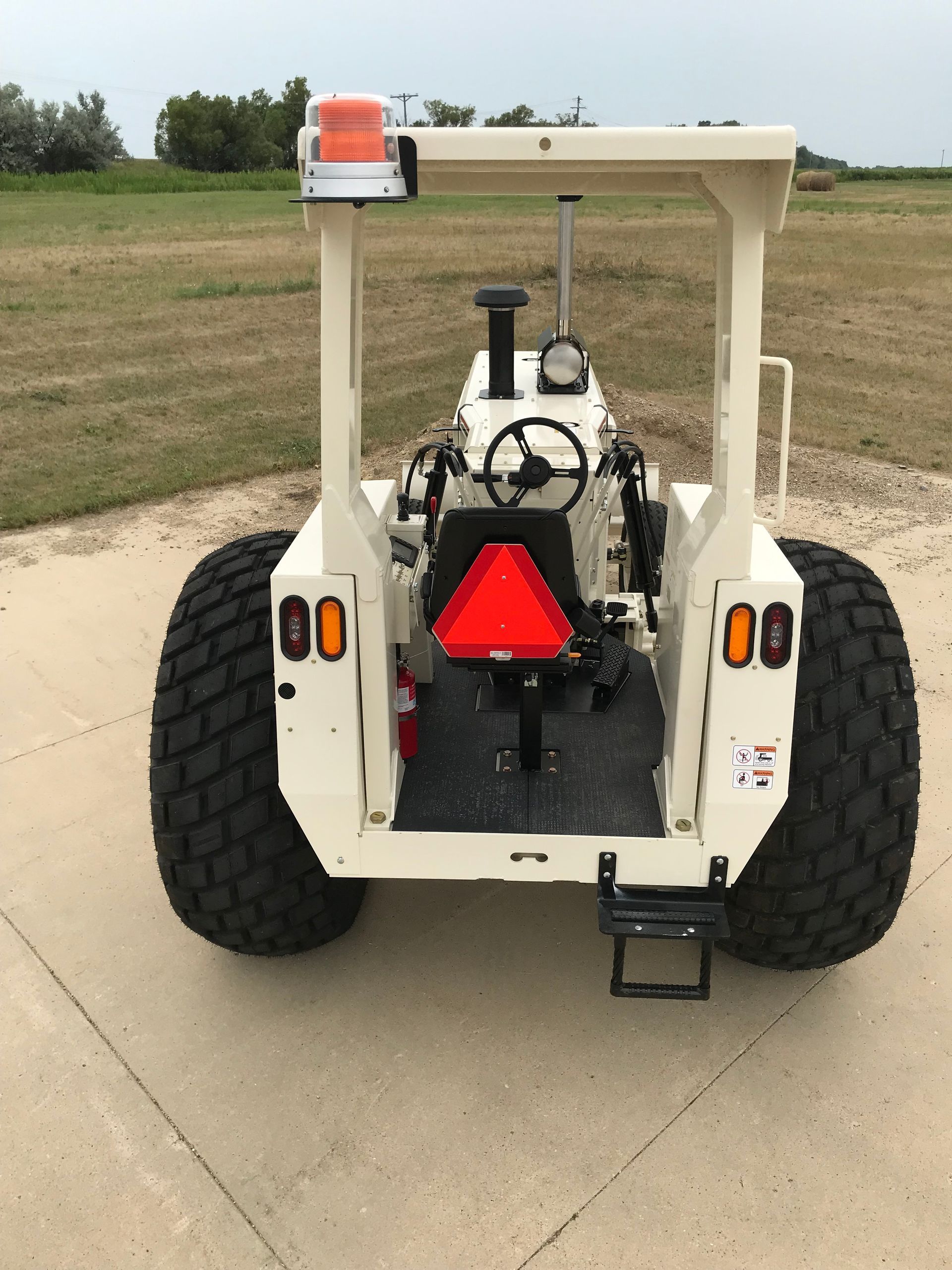 Rear view of a beige tractor with large black tires on a paved surface. It has a roll cage, warning light, and a slow-moving vehicle emblem.