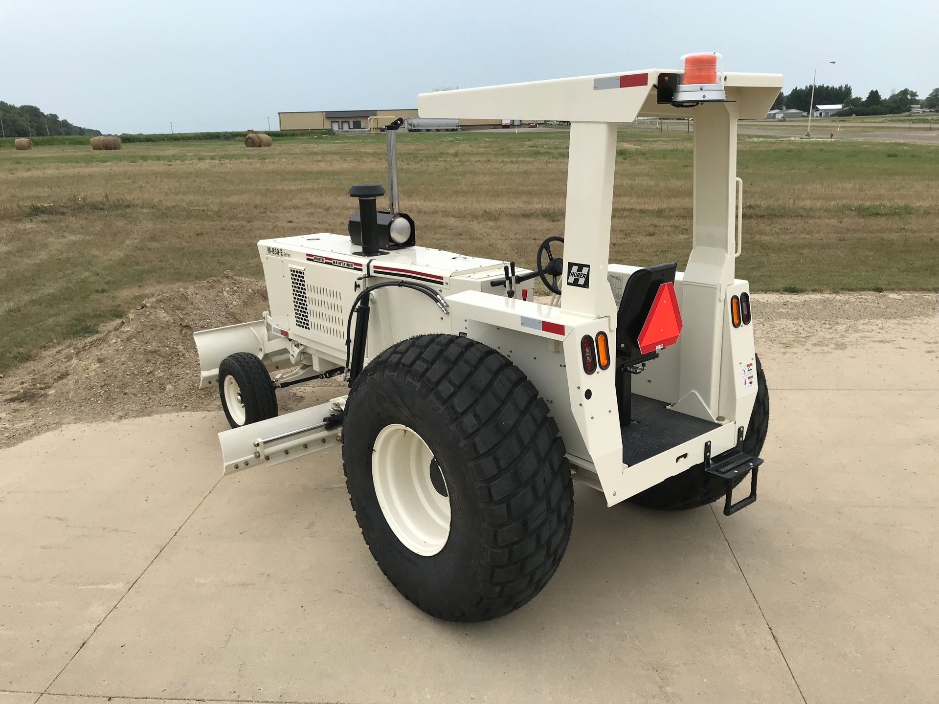White and black tractor on a concrete surface with large tires, in front of a field and overcast sky.