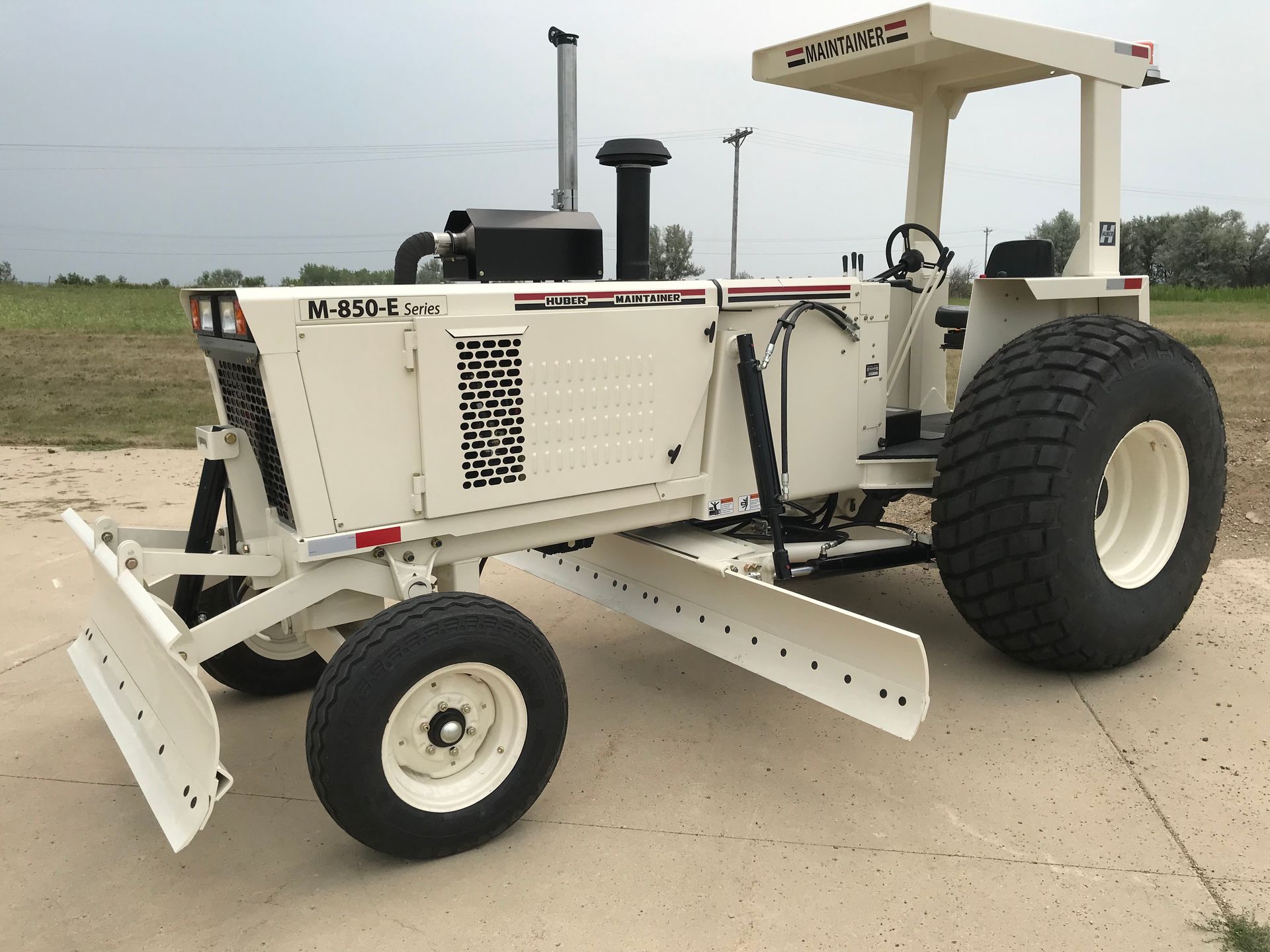 Cream-colored road grader on concrete, facing left, with a blade and large tires, under a cloudy sky.