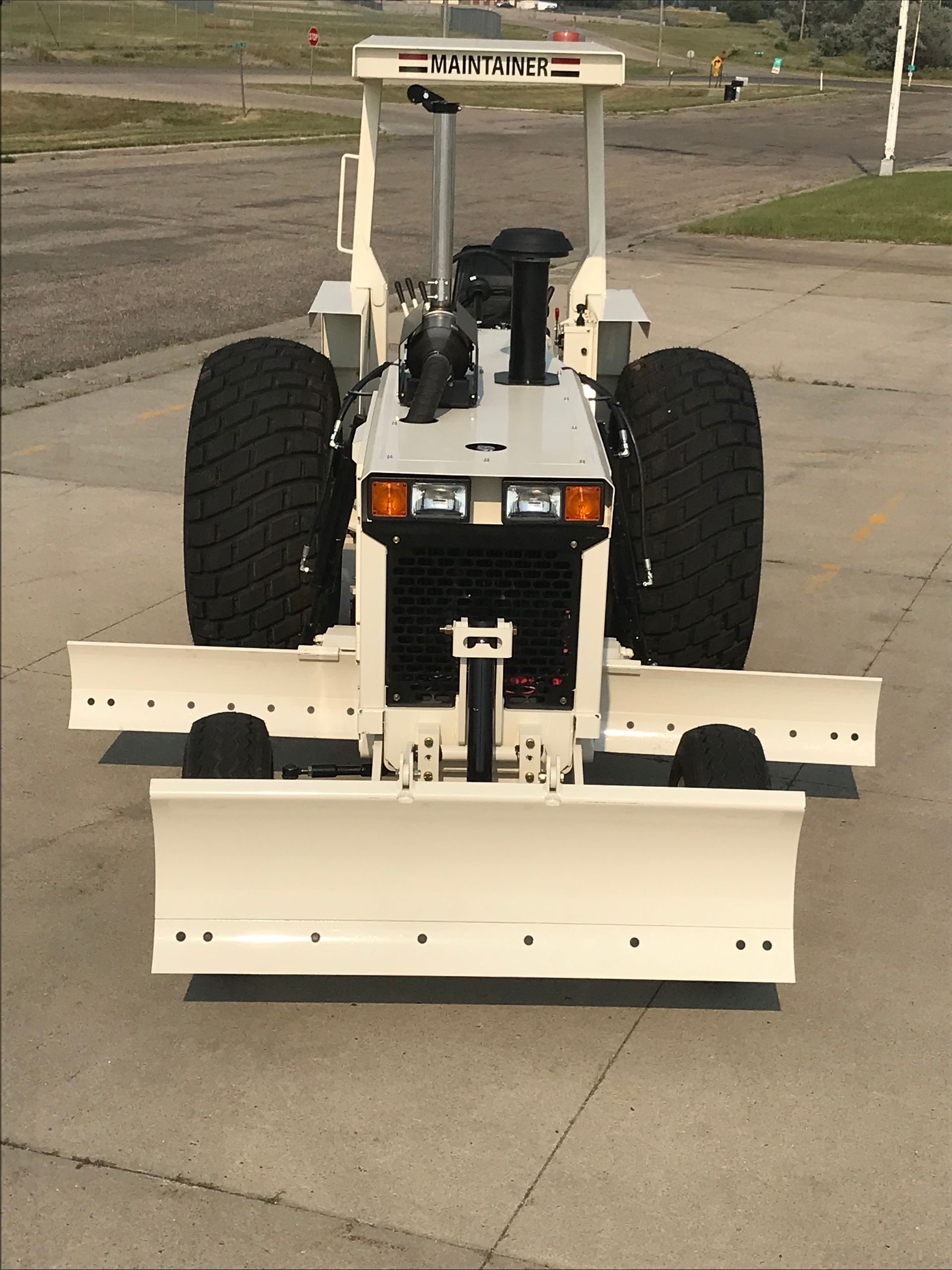 White tractor with a snowplow on pavement, under a cloudy sky.