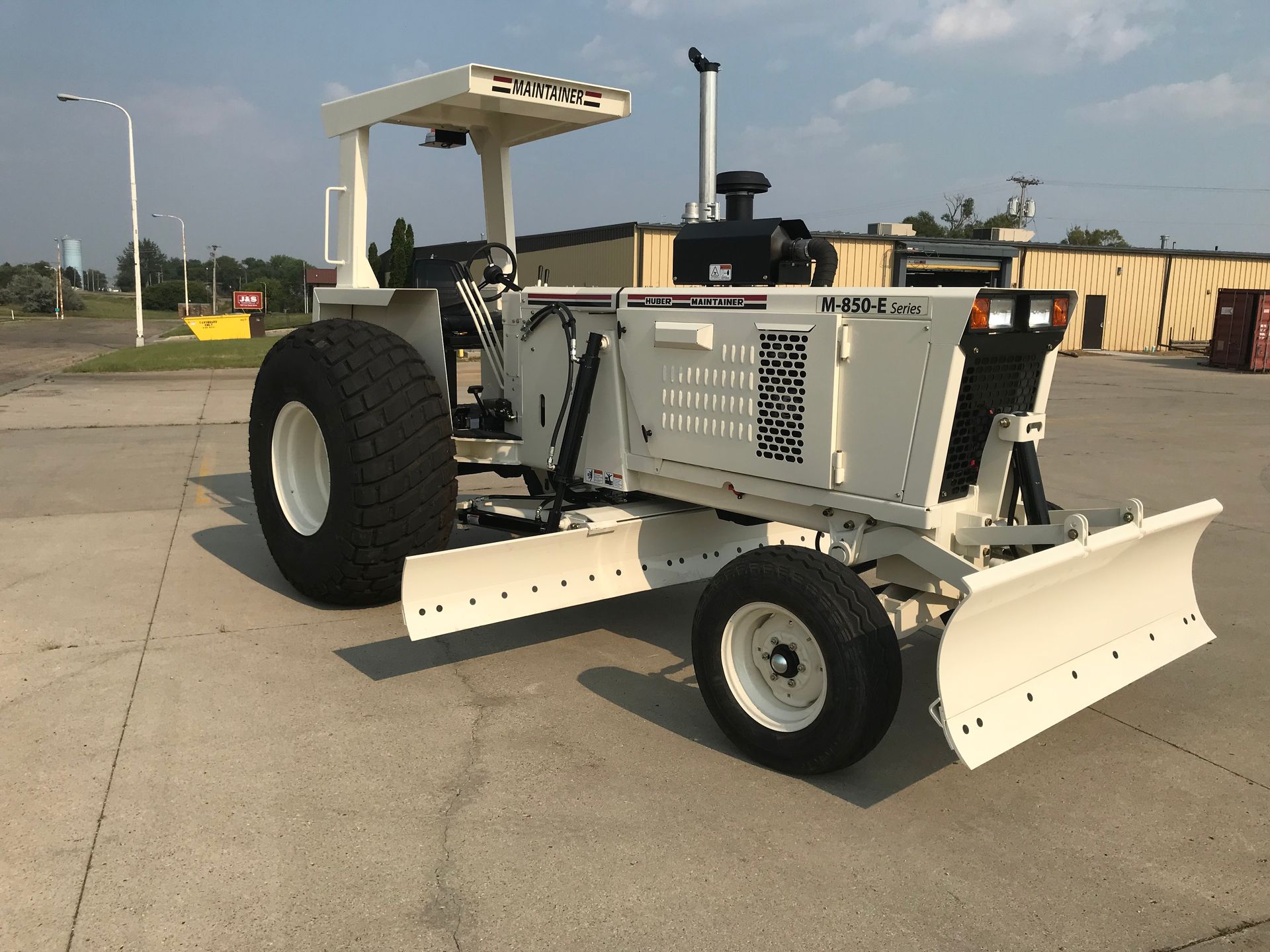 White grader with blade on pavement, under a blue sky.