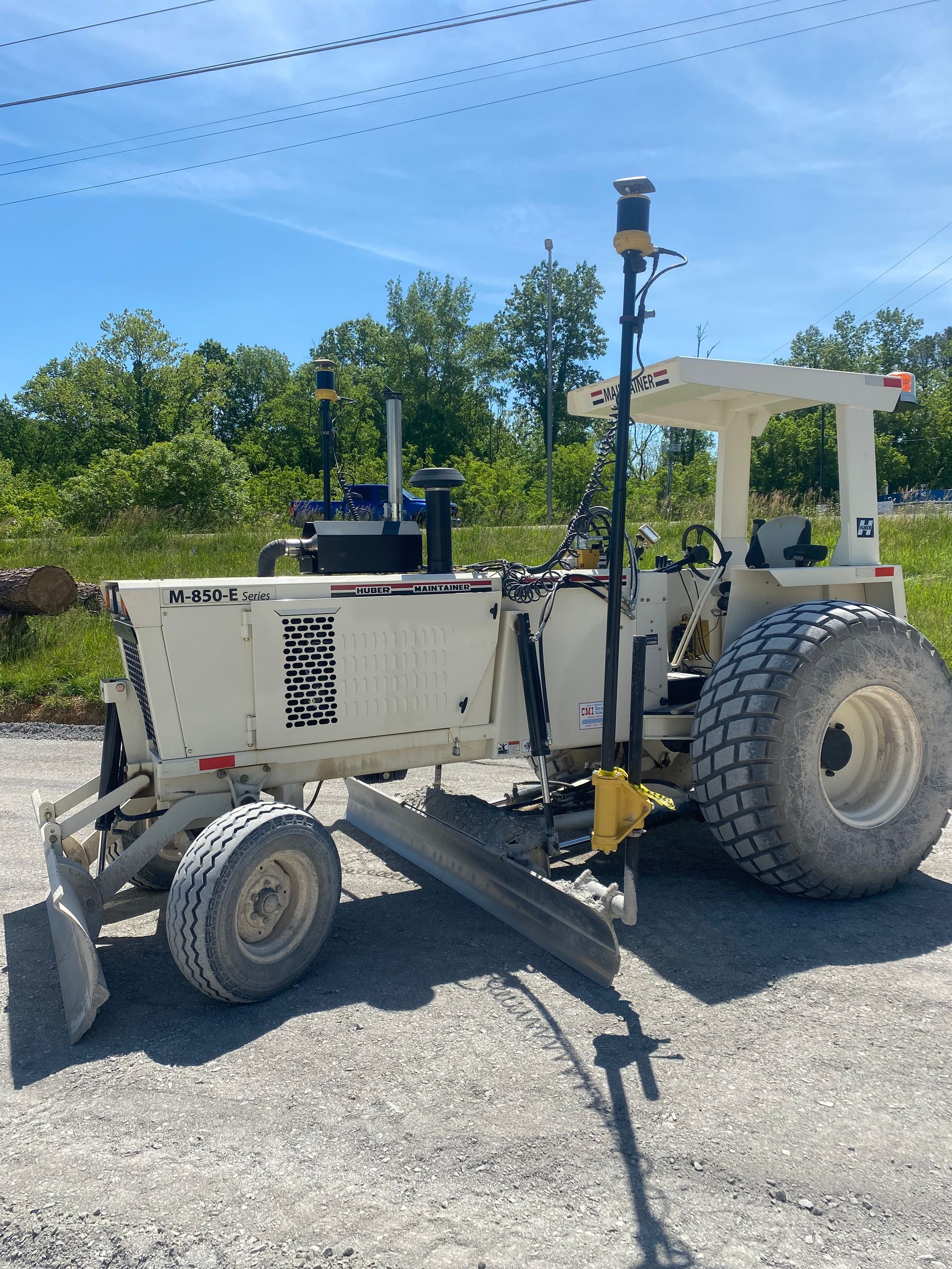 Cream-colored road grader with large tires and a GPS antenna on a gravel surface, against a backdrop of trees and a blue sky.