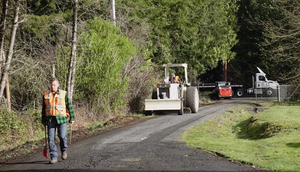 Person in safety vest walks on gravel road past a tractor and truck.