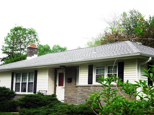 A white house with a gray roof and black shutters is surrounded by trees and bushes.
