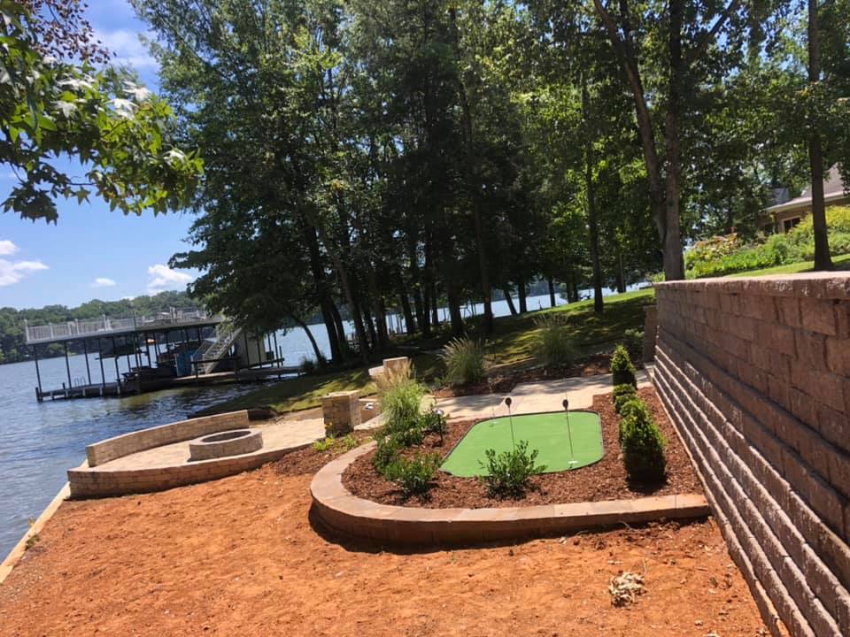 A small green golf course is surrounded by trees next to a lake.