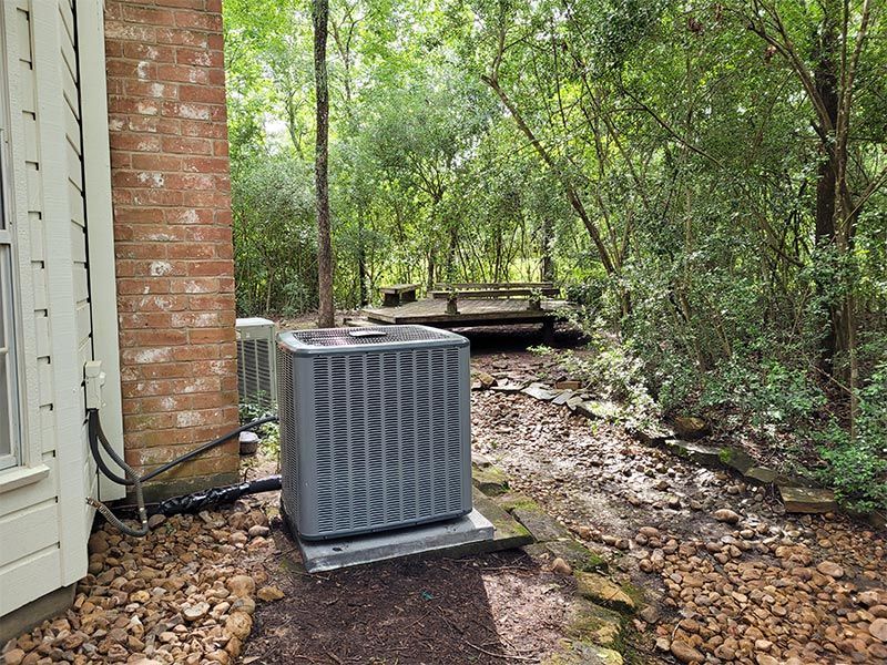 An air conditioner is sitting outside of a house in the woods.