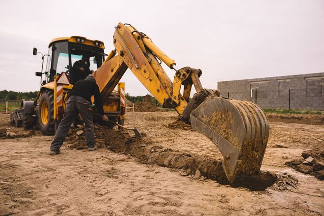 A worker uses a shovel near a yellow backhoe digging a trench at a construction site.