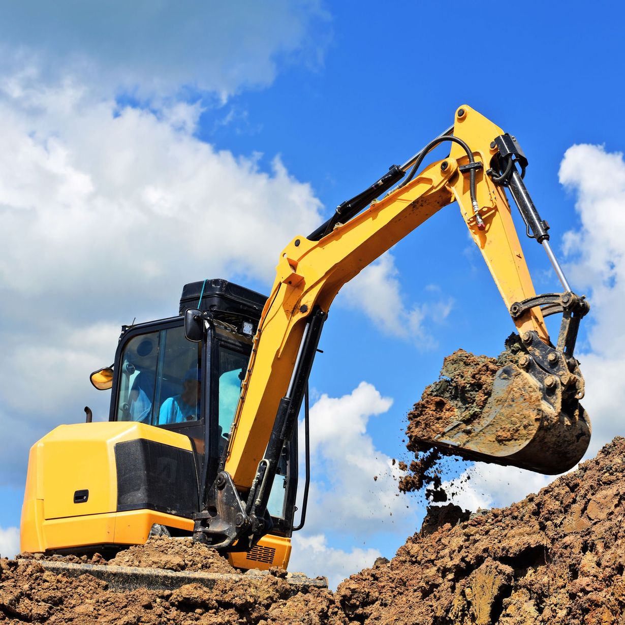 A yellow excavator digging into a pile of dirt against a bright blue sky with scattered white clouds.