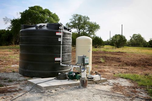 A large black water tank and a beige pressure tank connected to a pump on a concrete pad in a rural outdoor field.