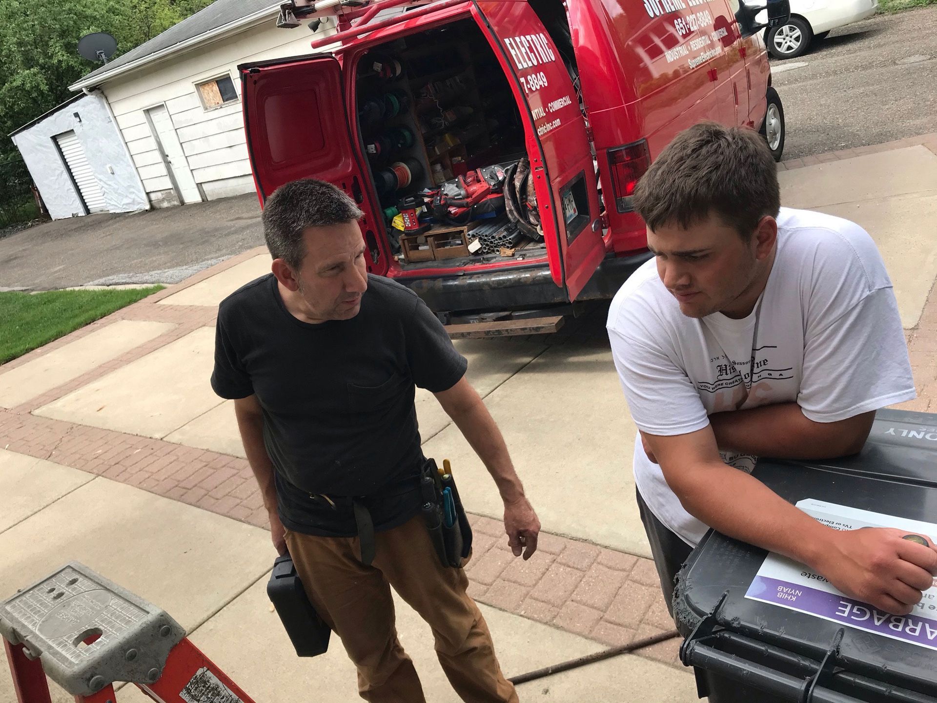 Two men beside a red van, one leaning on a car hood in a driveway or street.