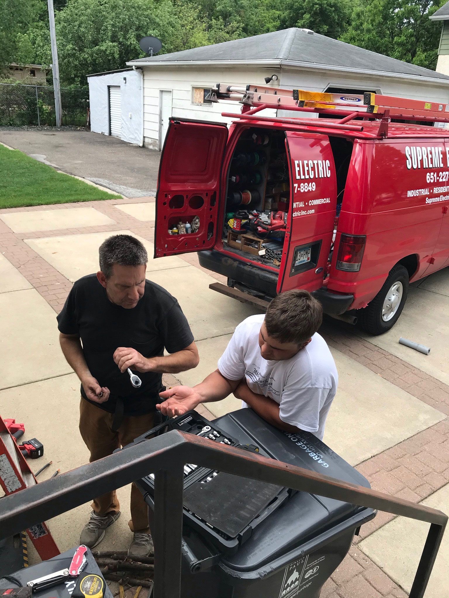 Two people work on an open black bin beside a red utility van in a driveway.