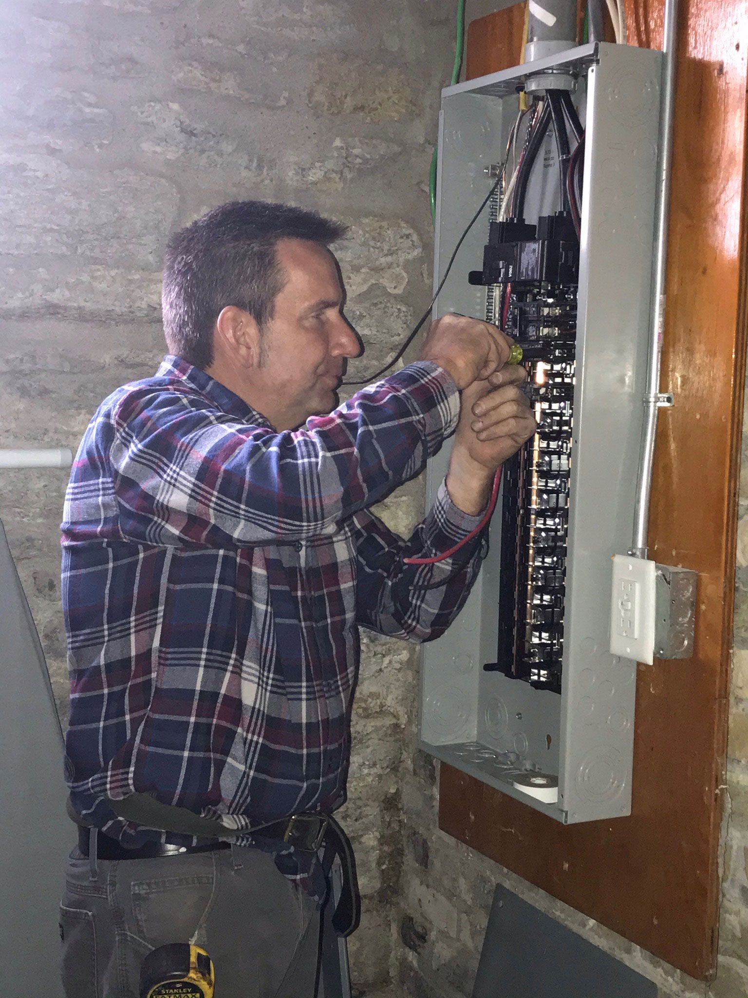 Man in plaid shirt working inside an open electrical breaker panel in a basement