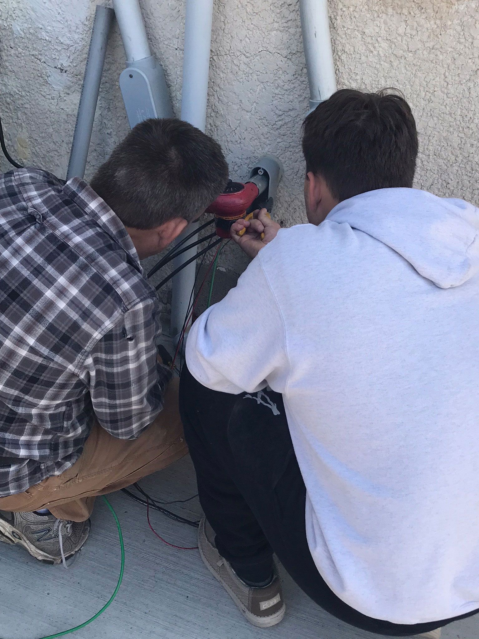 Two people crouch beside a wall, working on exposed cables with a red power tool.