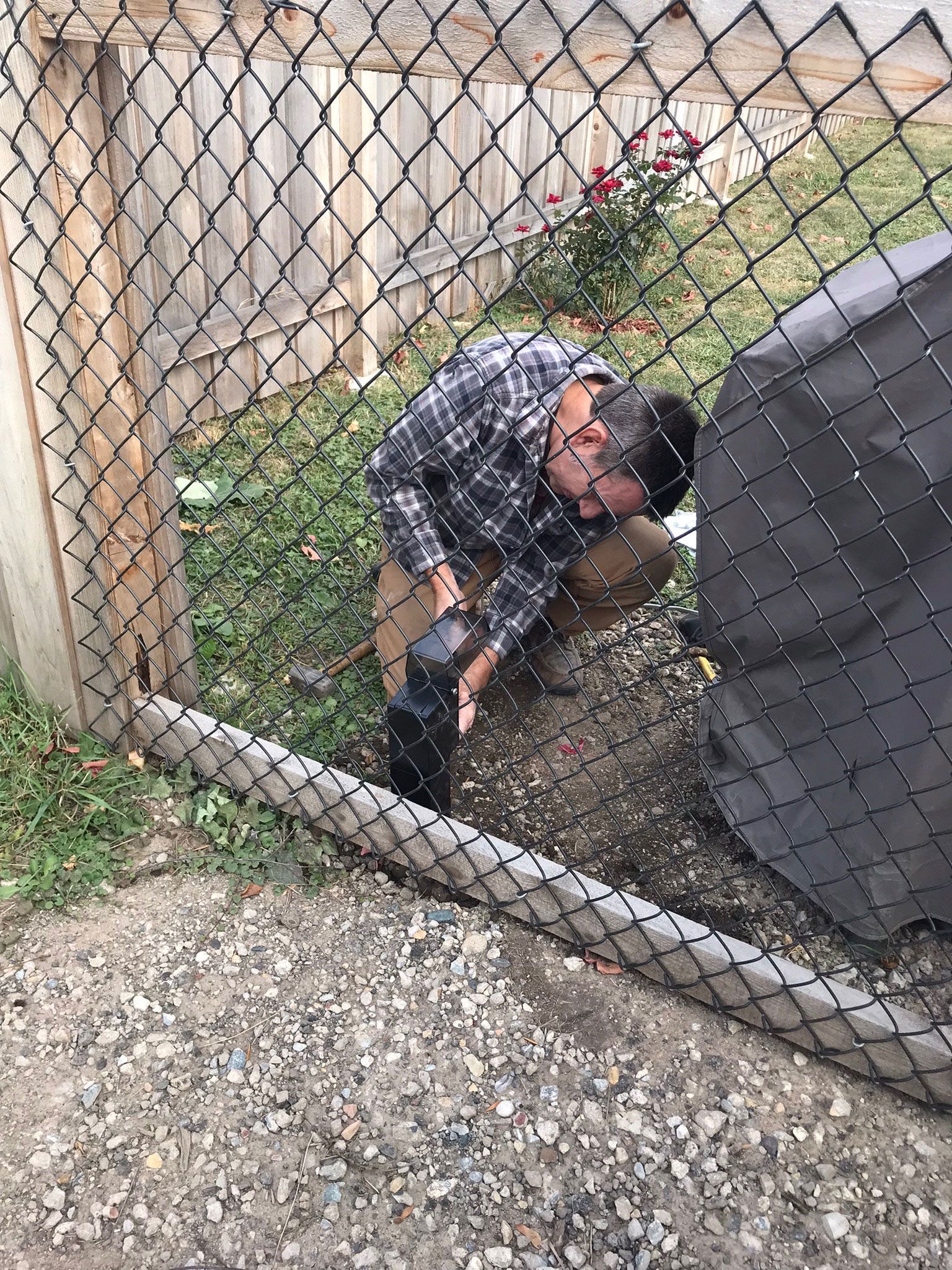 Man crouching by a chain-link fence beside a gray structure, inspecting the ground