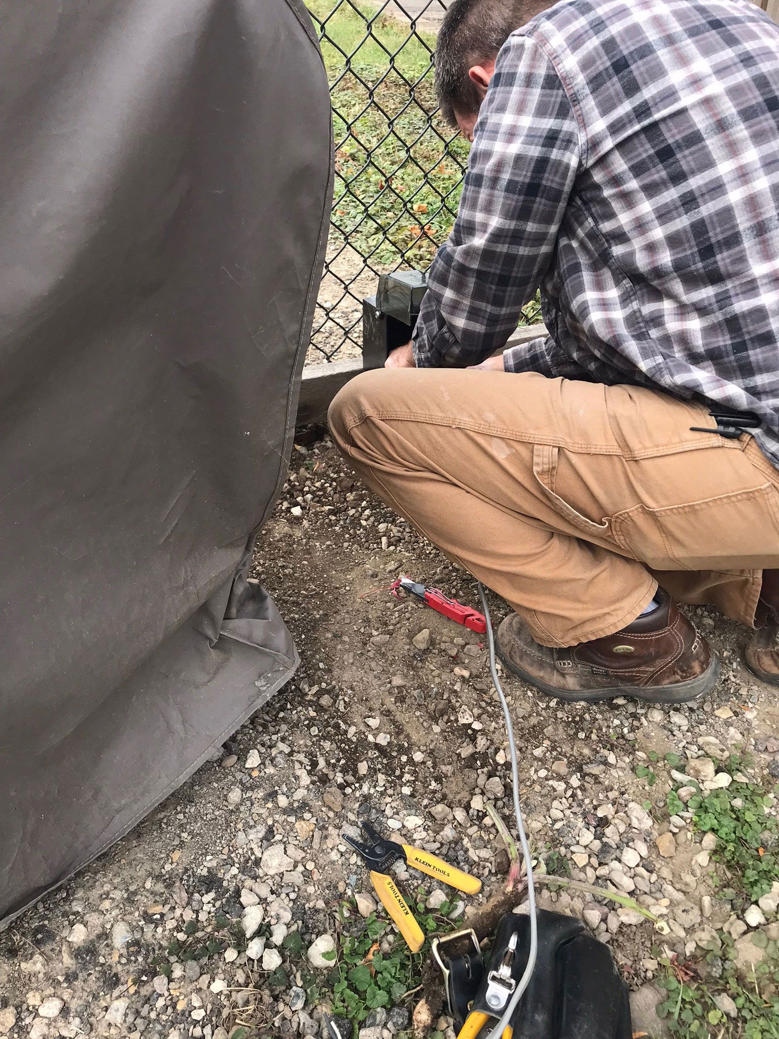 Two people crouch by a chain-link fence on gravel, with a yellow tool and black bag on the ground.