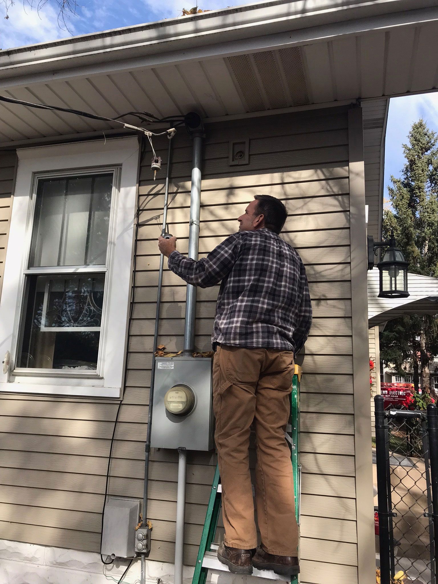 Person on ladder inspecting roof gutter beside gray house with utility meter.