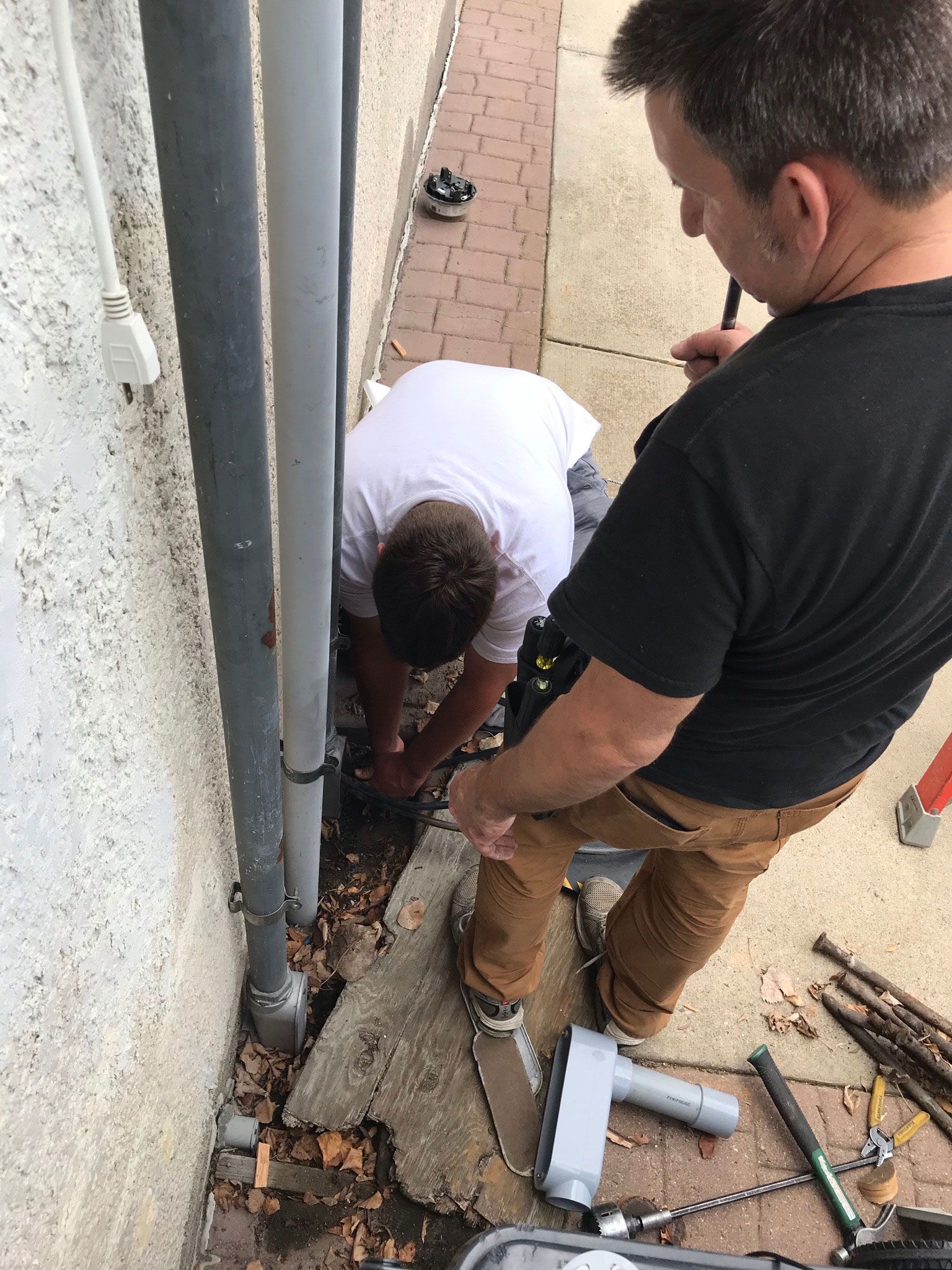 Two workers repair a damaged wall base beside a building, using tools and concrete debris.
