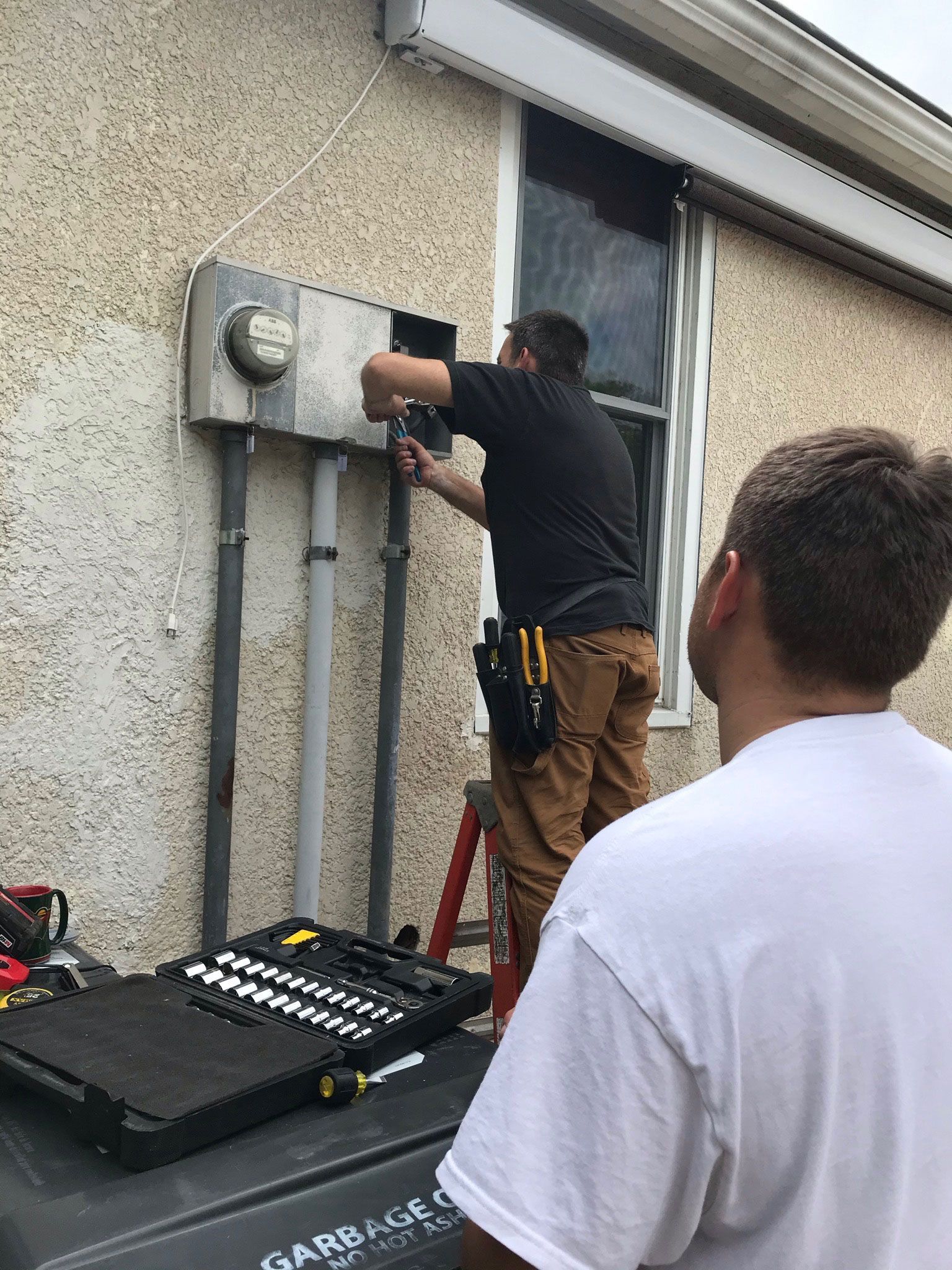 Technician using a drill beside an exterior wall-mounted electrical box, with tools on the ground