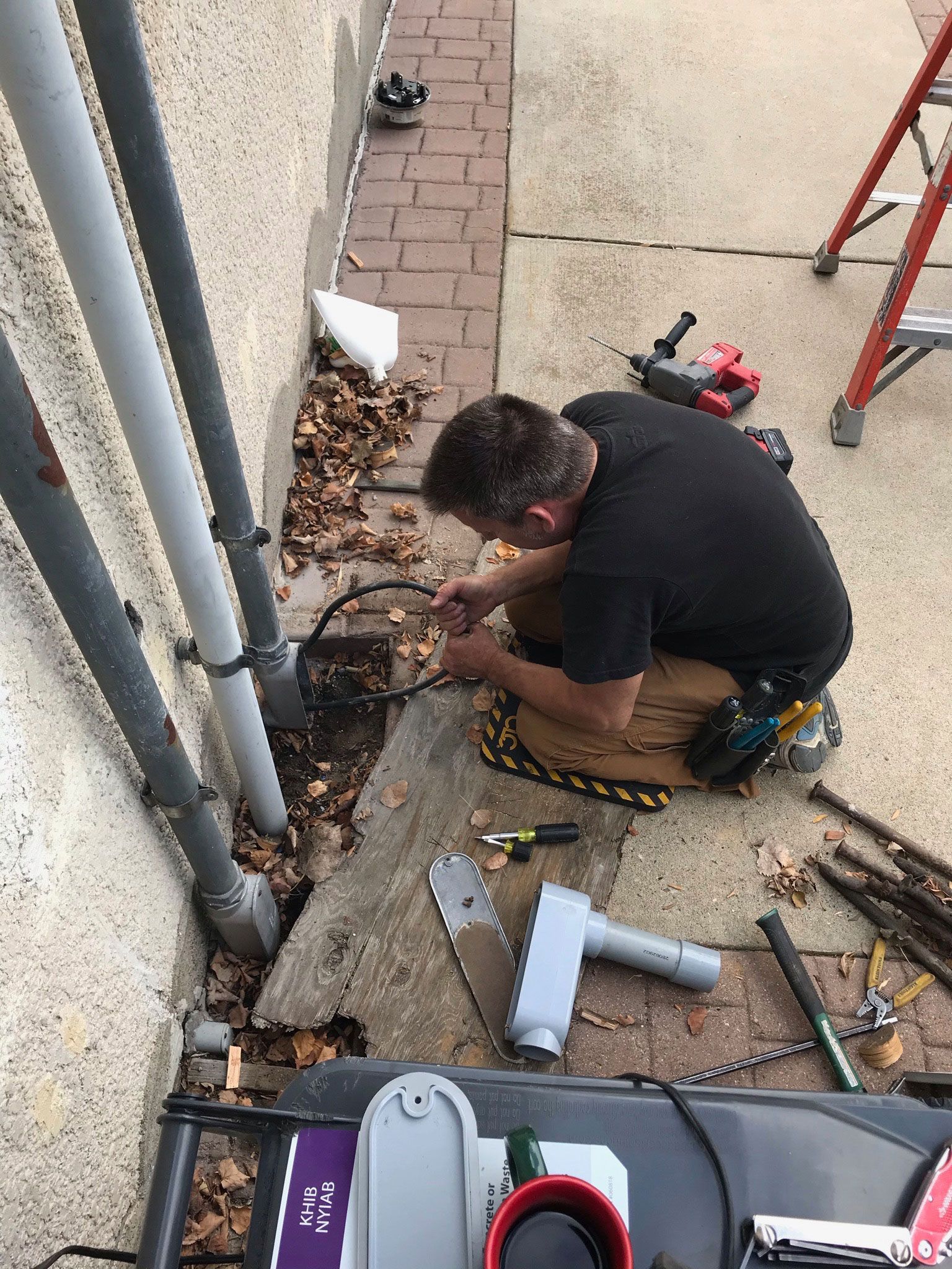Person kneeling by wall, using tools to work on a cable or pipe beside a walkway.