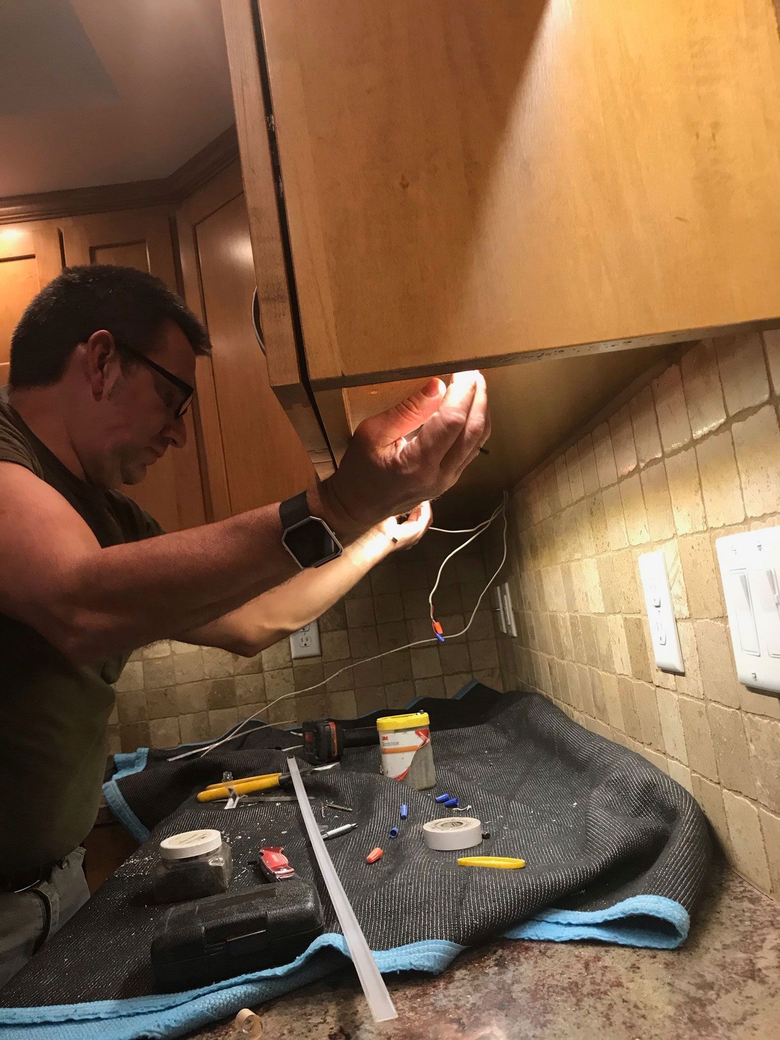 Person working under a kitchen cabinet with tools and exposed wires on the counter.