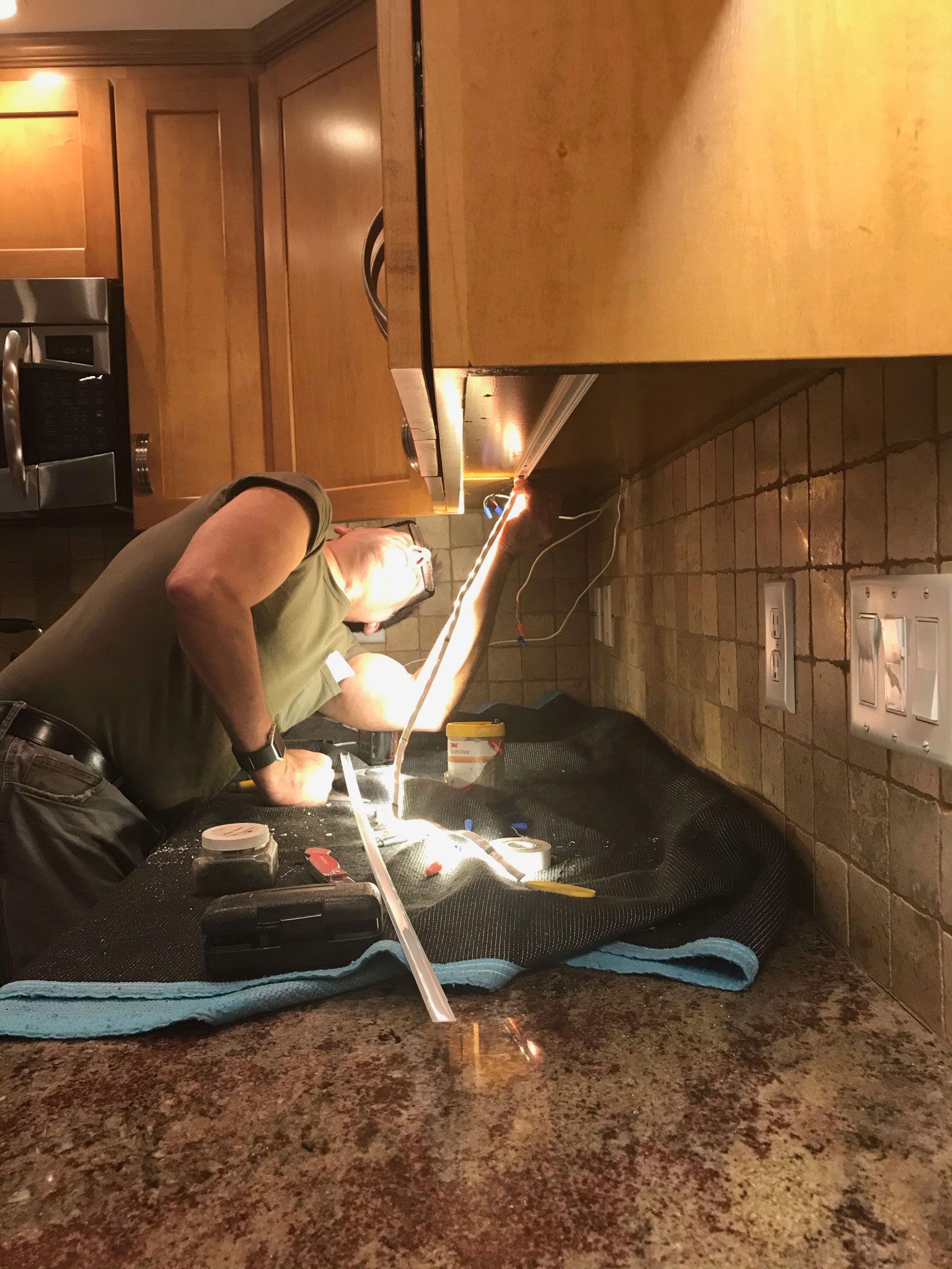 Person fixing kitchen sink under cabinet light, with tools on countertop and backsplash tiles nearby