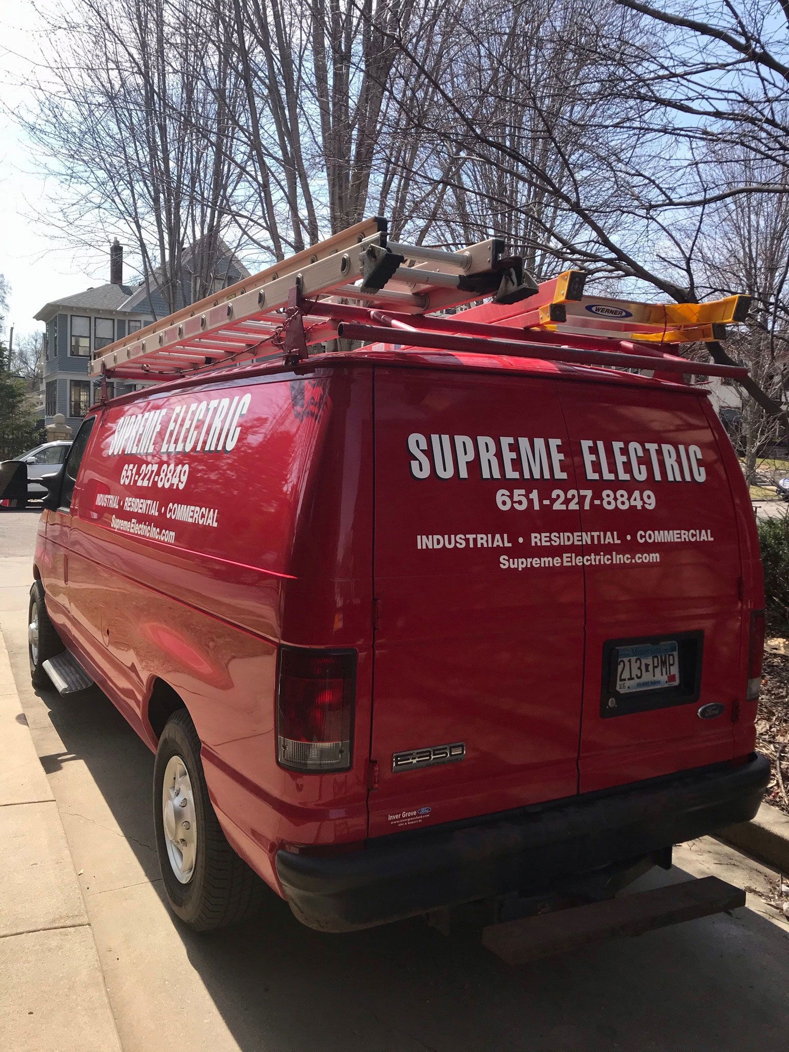 Red van with ladders on roof, labeled “Supreme Electric,” parked on a street by trees.