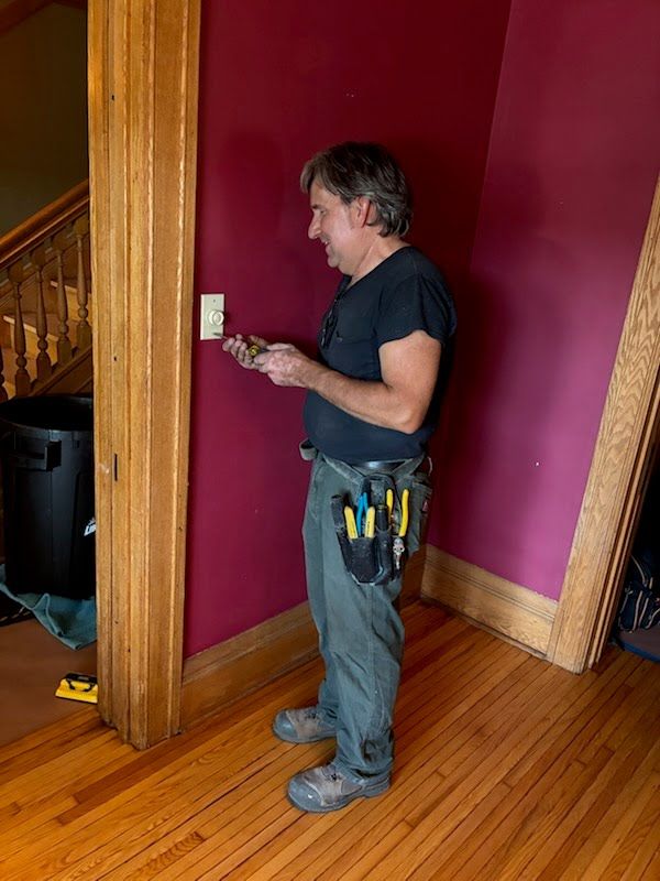 Man standing by a light switch in a pink-walled hallway, holding tools in his belt