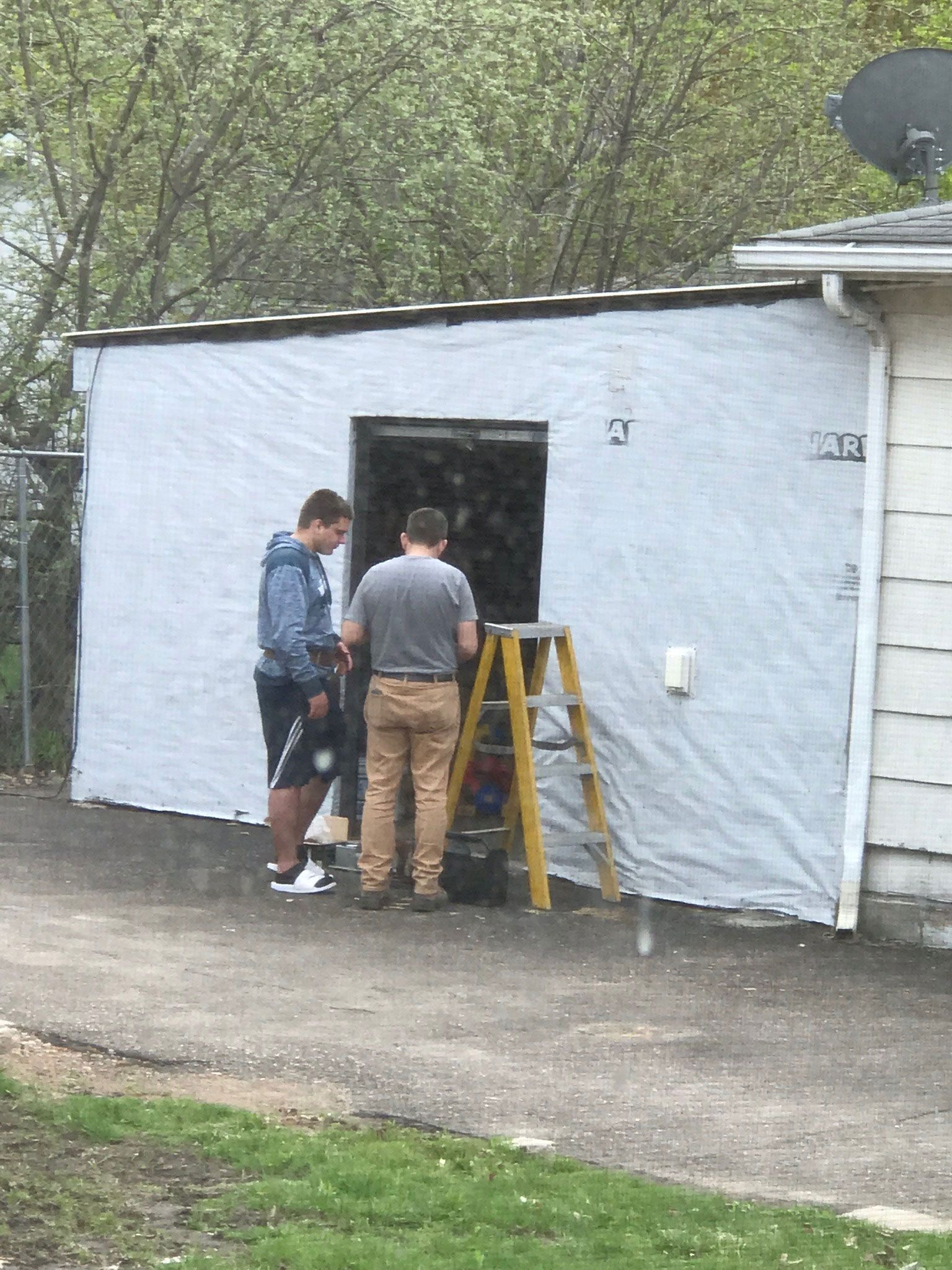 Two people inspect a small white building entrance beside a yellow ladder outdoors.