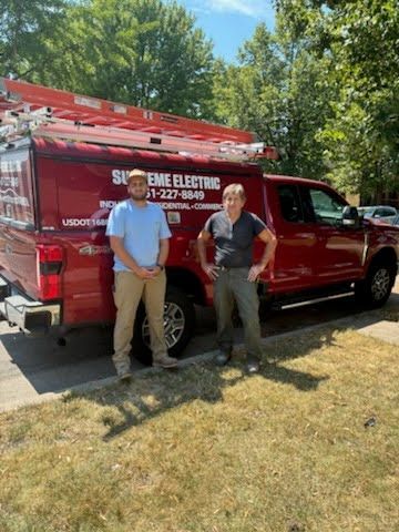 Two people standing beside a red utility truck in a sunny wooded area