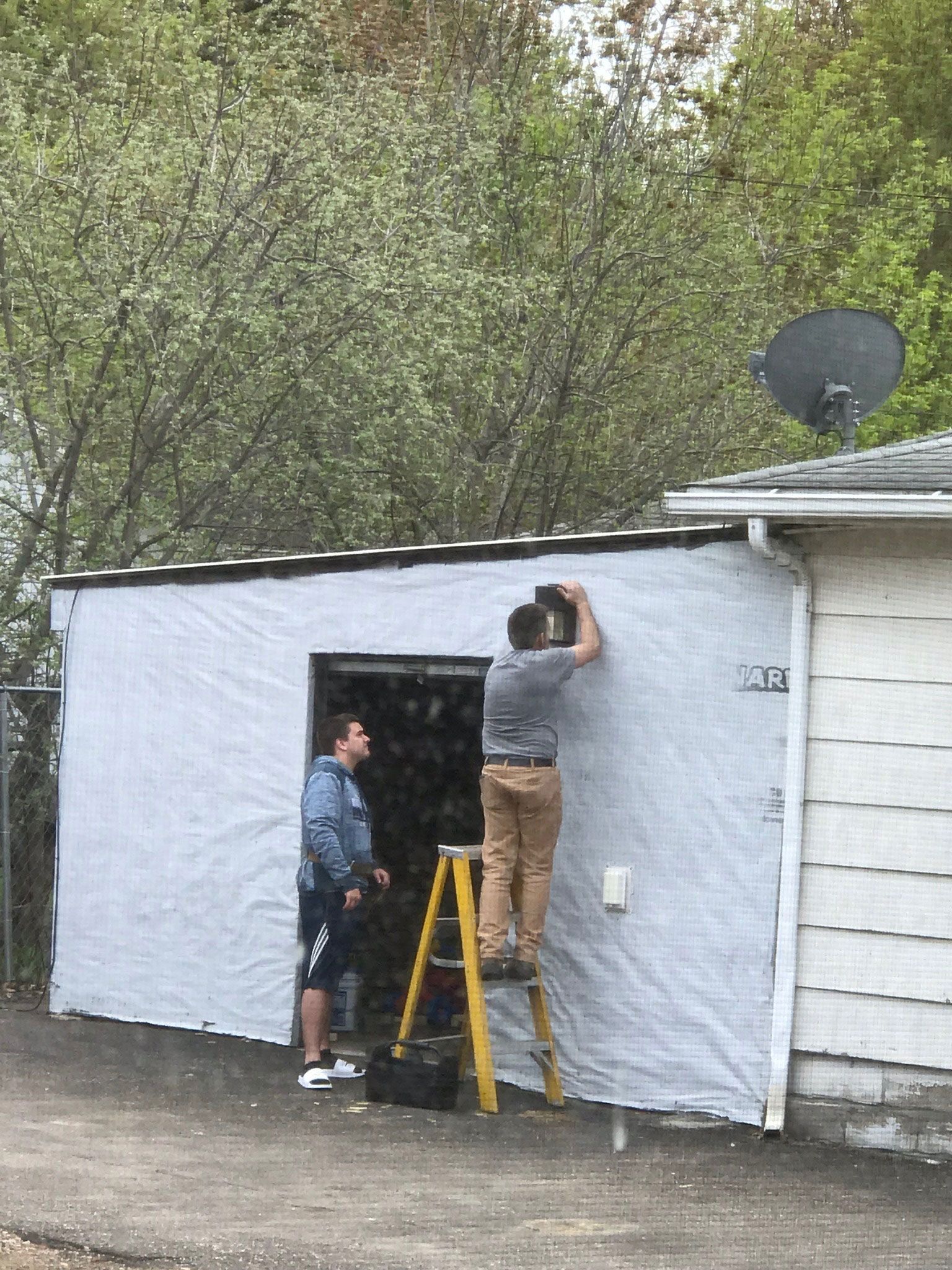 Two people working on a tarp-covered wall beside a house, with a ladder and satellite dish visible.