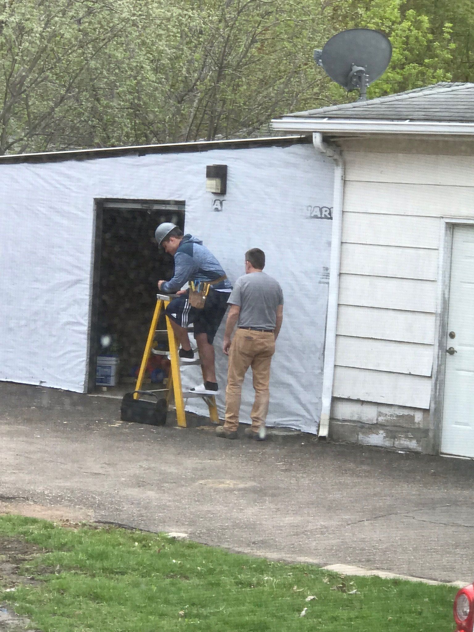 Two workers inspecting a white garage wall beside a doorway, one on a step ladder outdoors.
