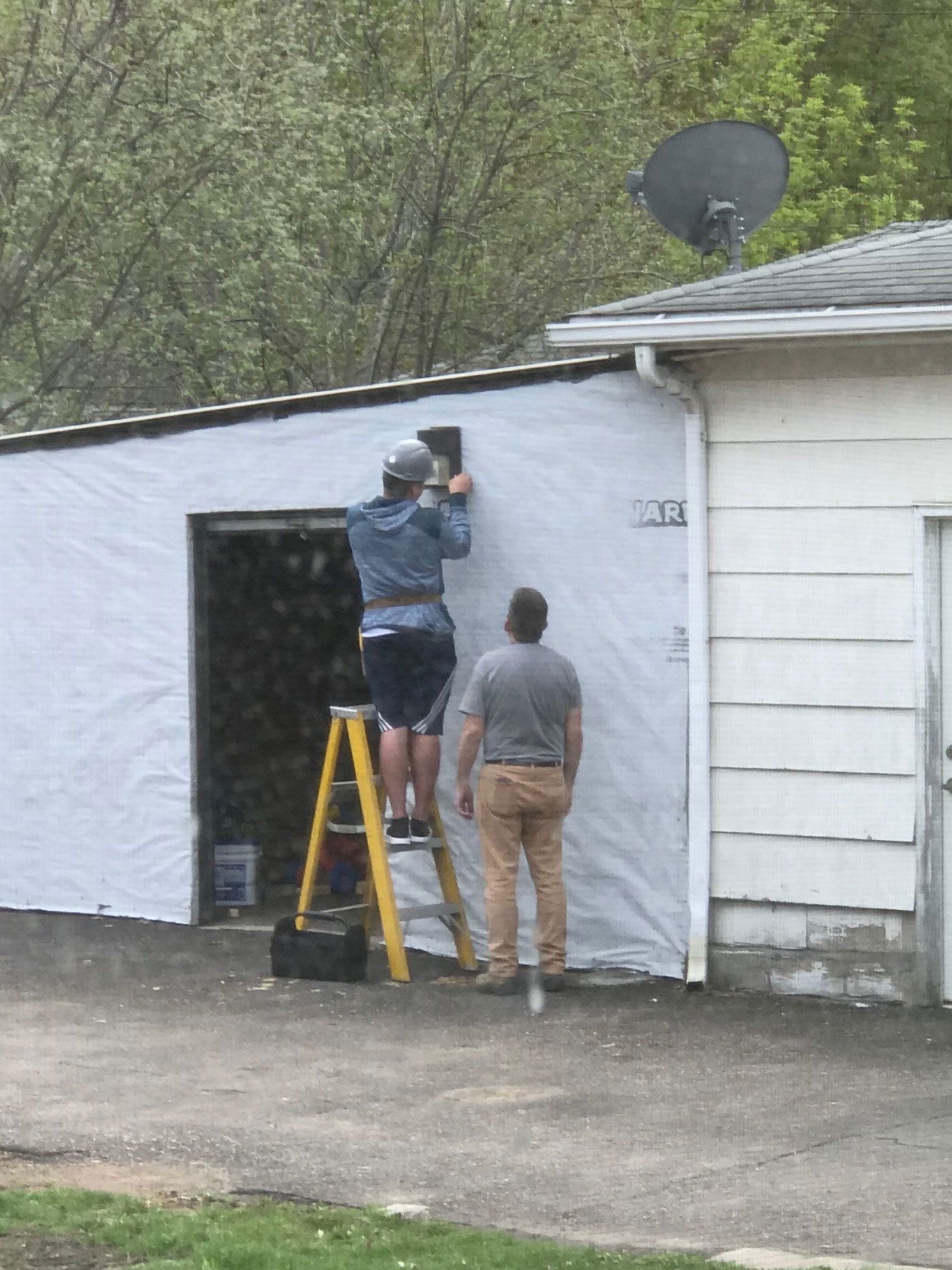 Two people work on a white garage wall, one on a yellow ladder under a satellite dish.