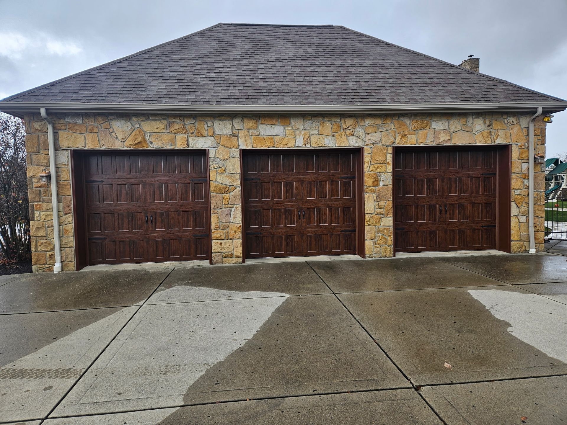 Three-car garage with brown doors and a stone exterior under a tiled roof, wet concrete driveway.