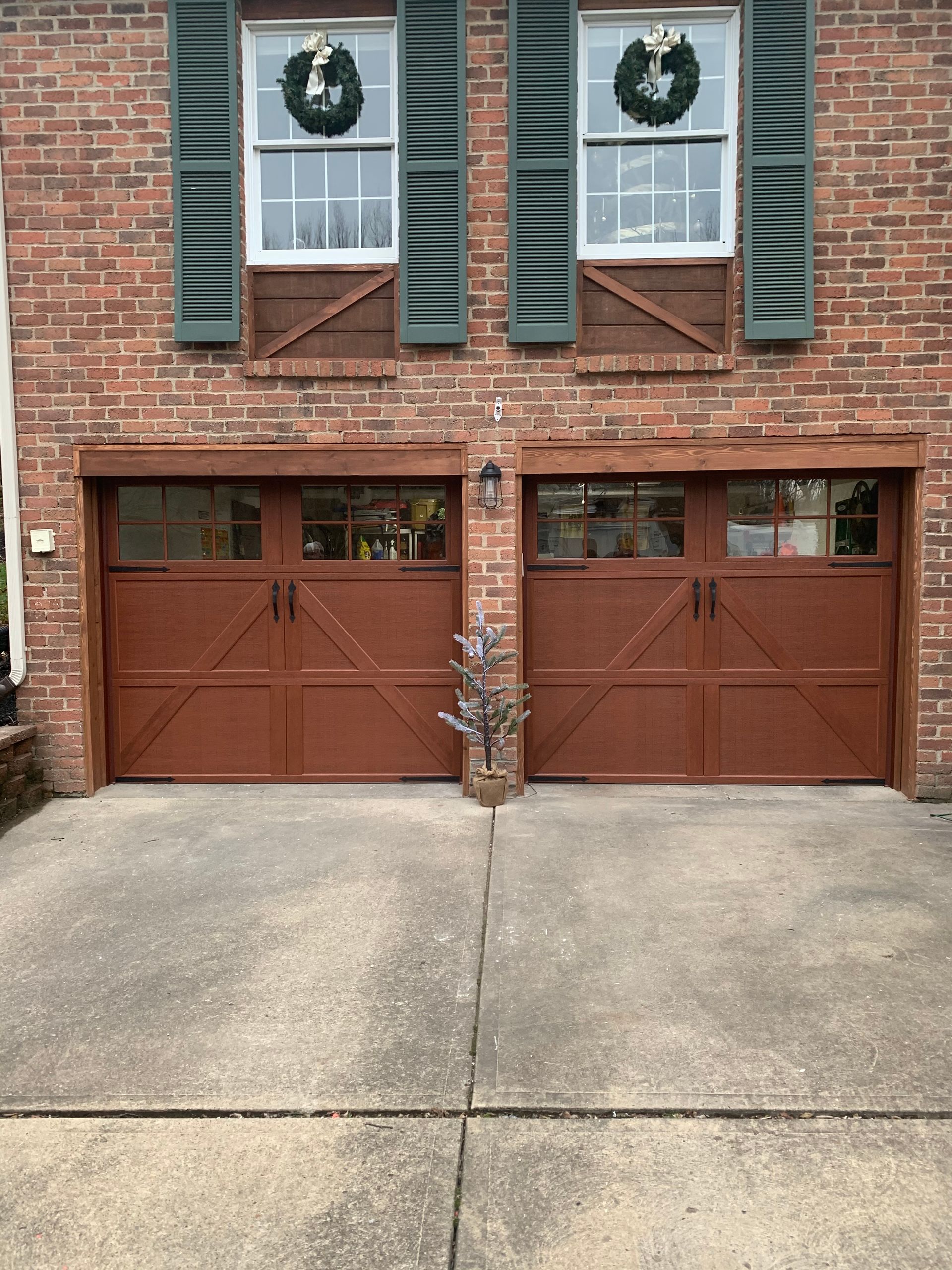 Two brown garage doors on a brick house, adorned with windows and Christmas wreaths.