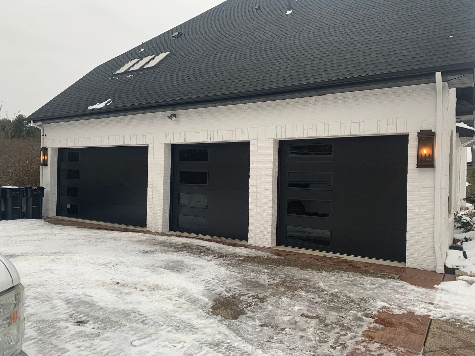 Three black garage doors on a white brick building with a dark roof, snow on the ground.