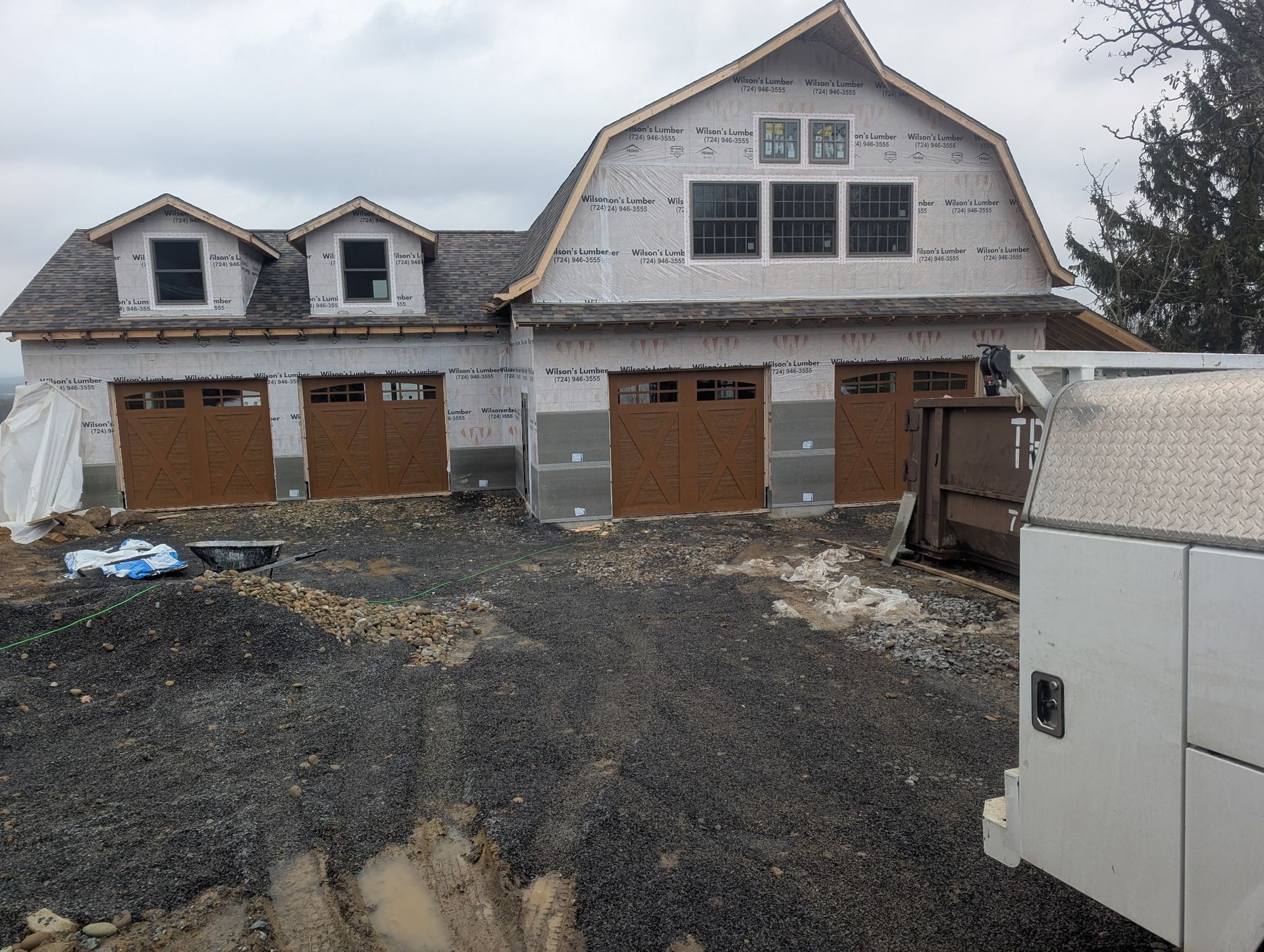 A barn-style building under construction with three garage doors and dormers, on a gravel lot.
