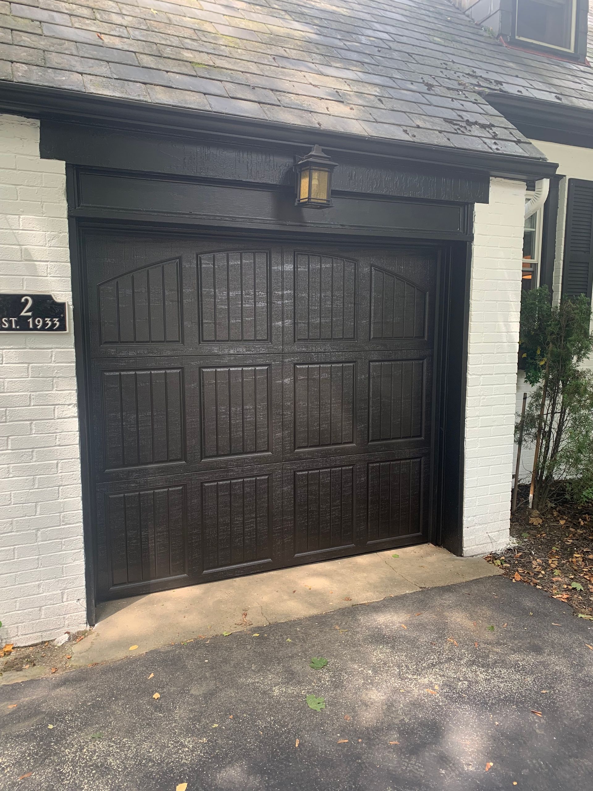 Black garage door with a light above, bordered by white brick and a driveway.