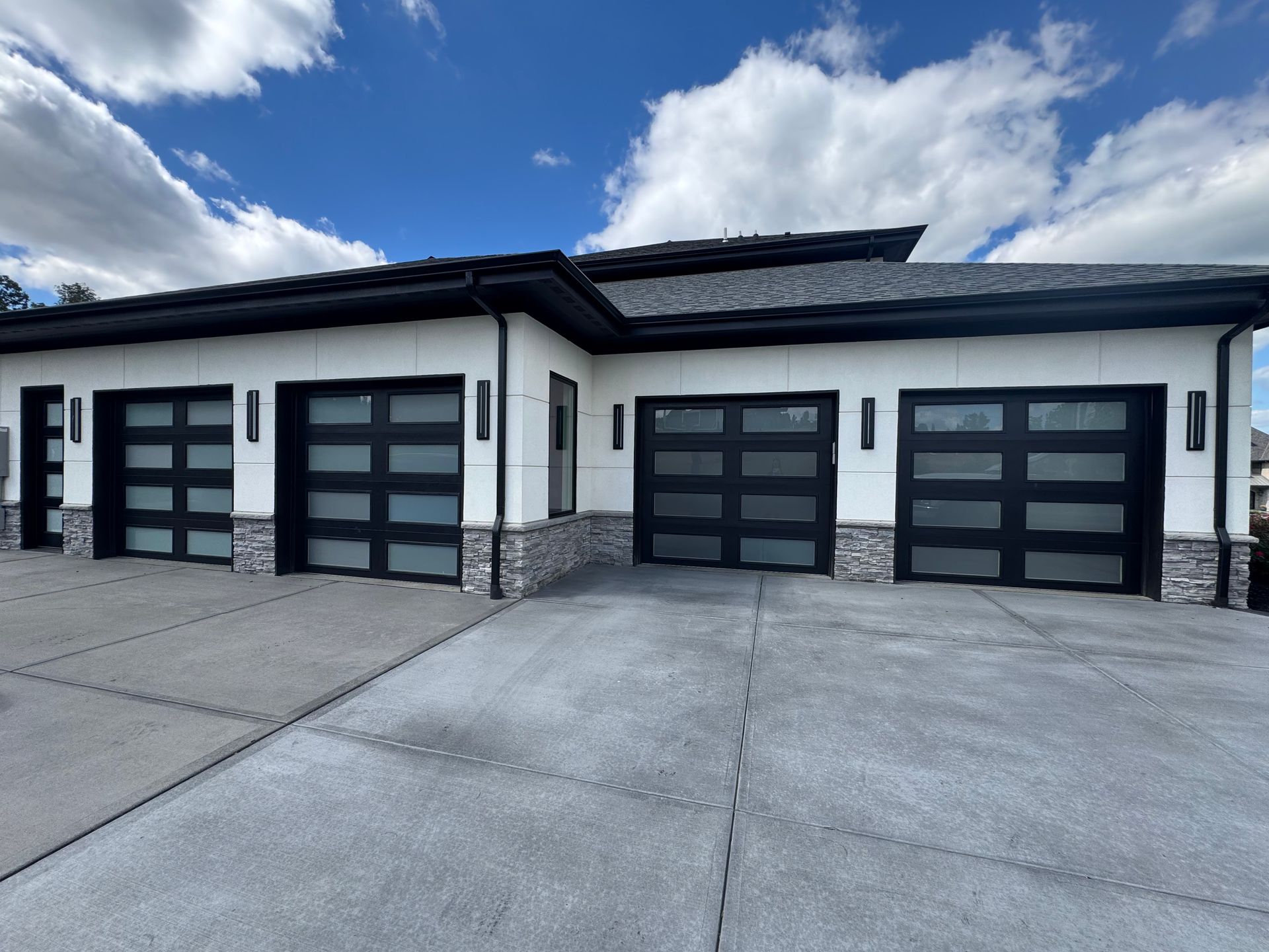 Three-car garage with black framed glass doors, light exterior, and blue sky. Concrete driveway.