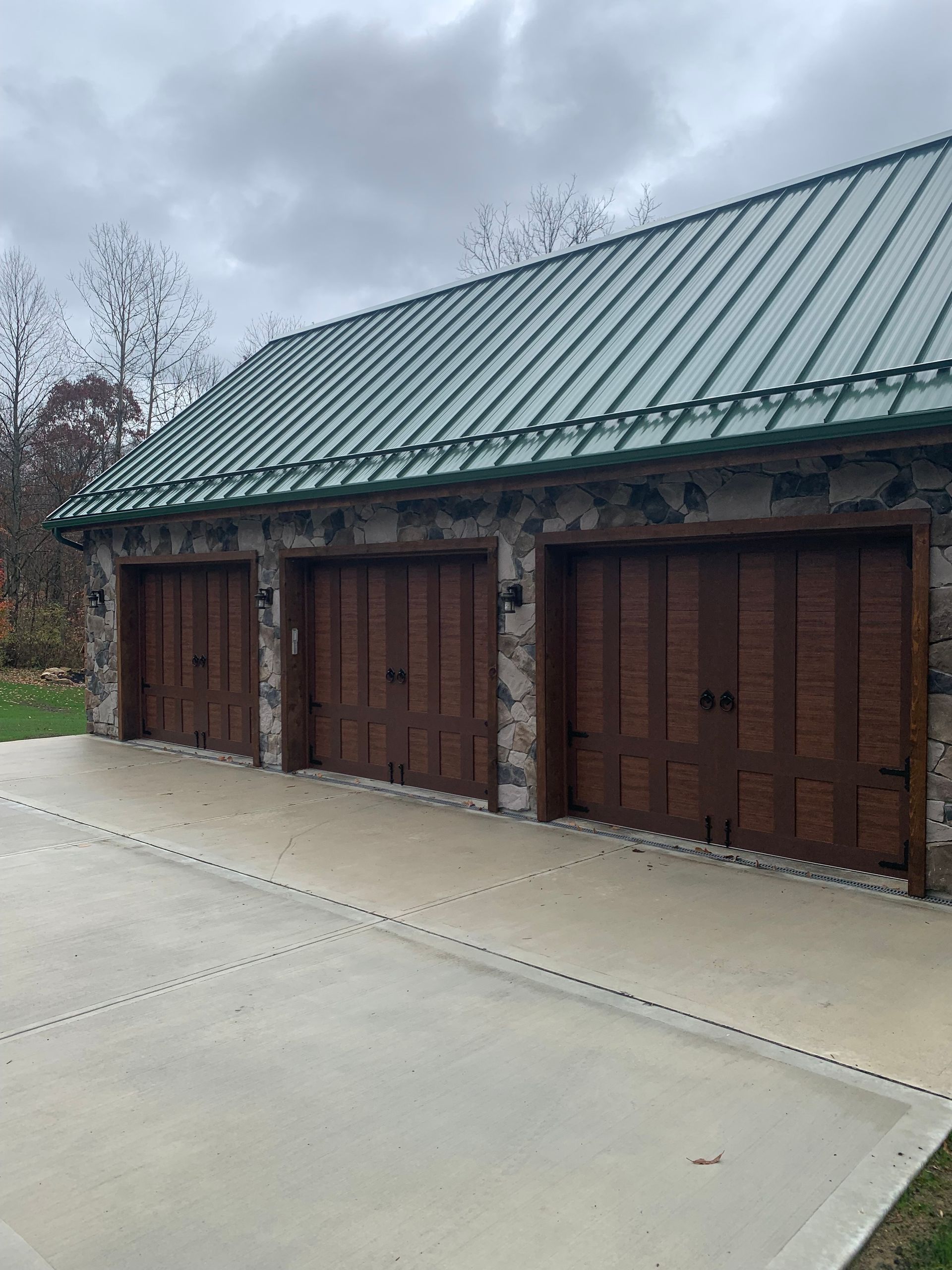 Three-bay garage with brown wooden doors and stone exterior under a green metal roof.