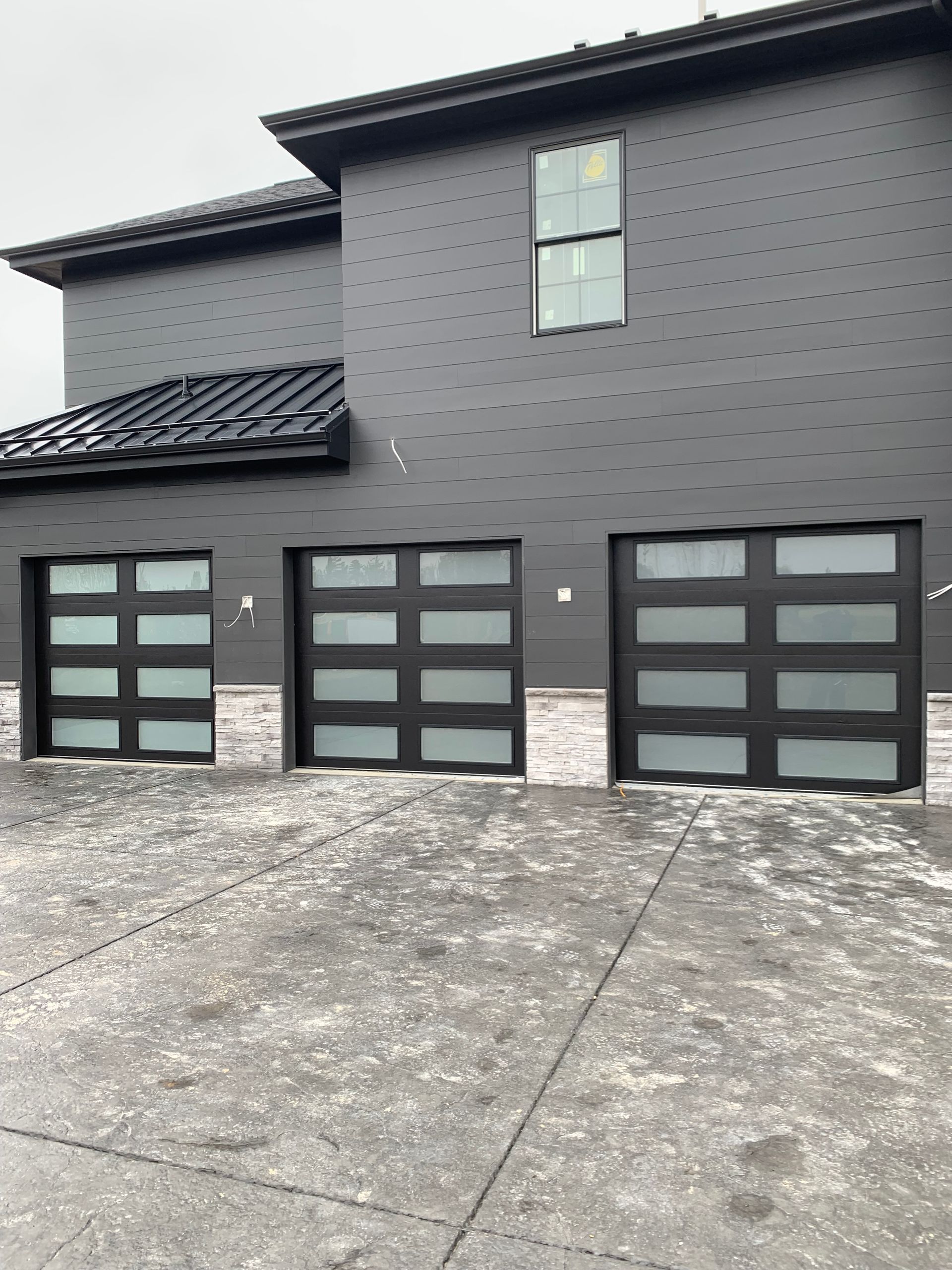Black modern home with three garage doors, frosted glass panels. Snow on the driveway.