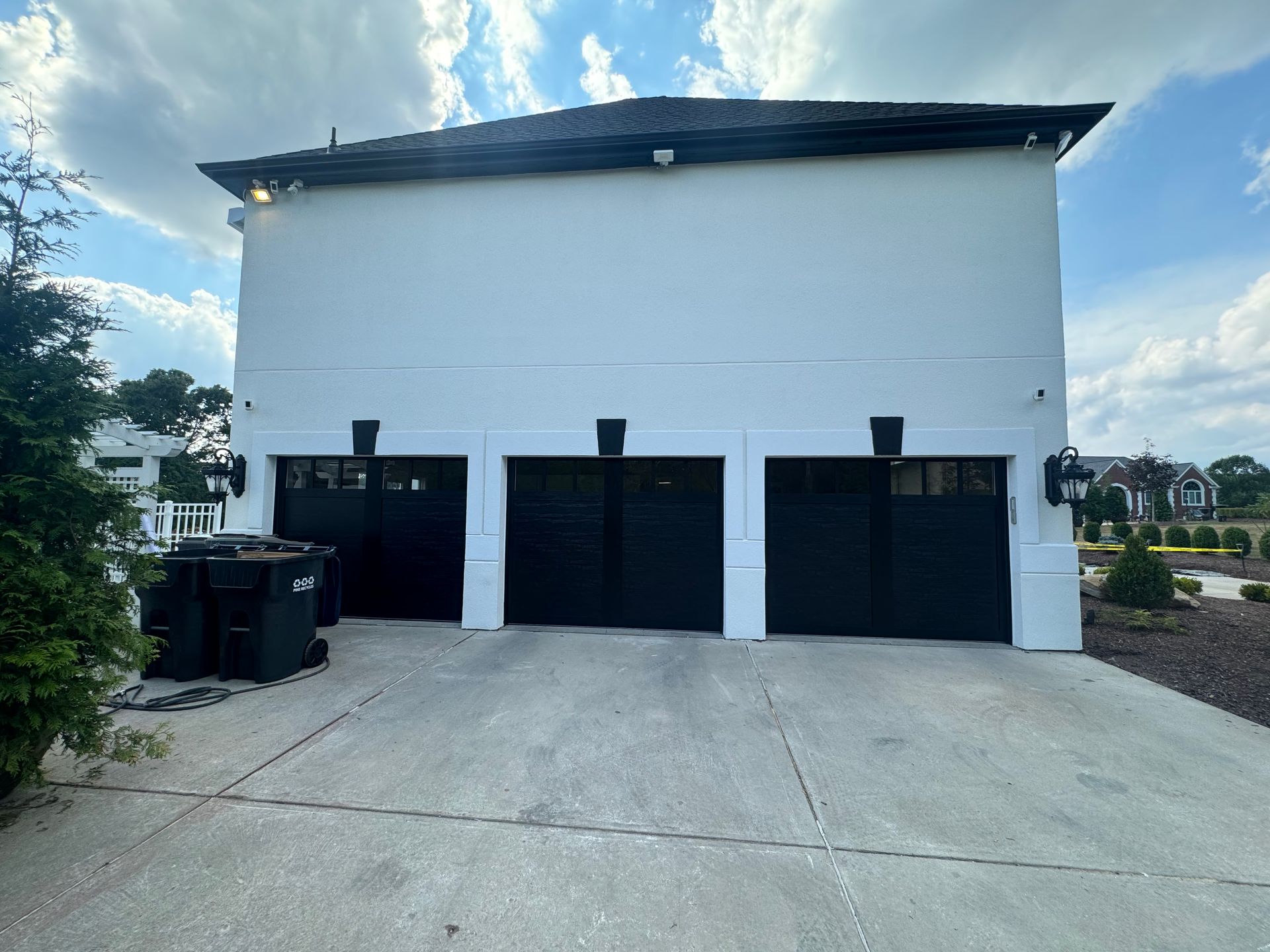 Three-car garage with black doors and white walls; a concrete driveway, two trash cans, and outdoor lights.