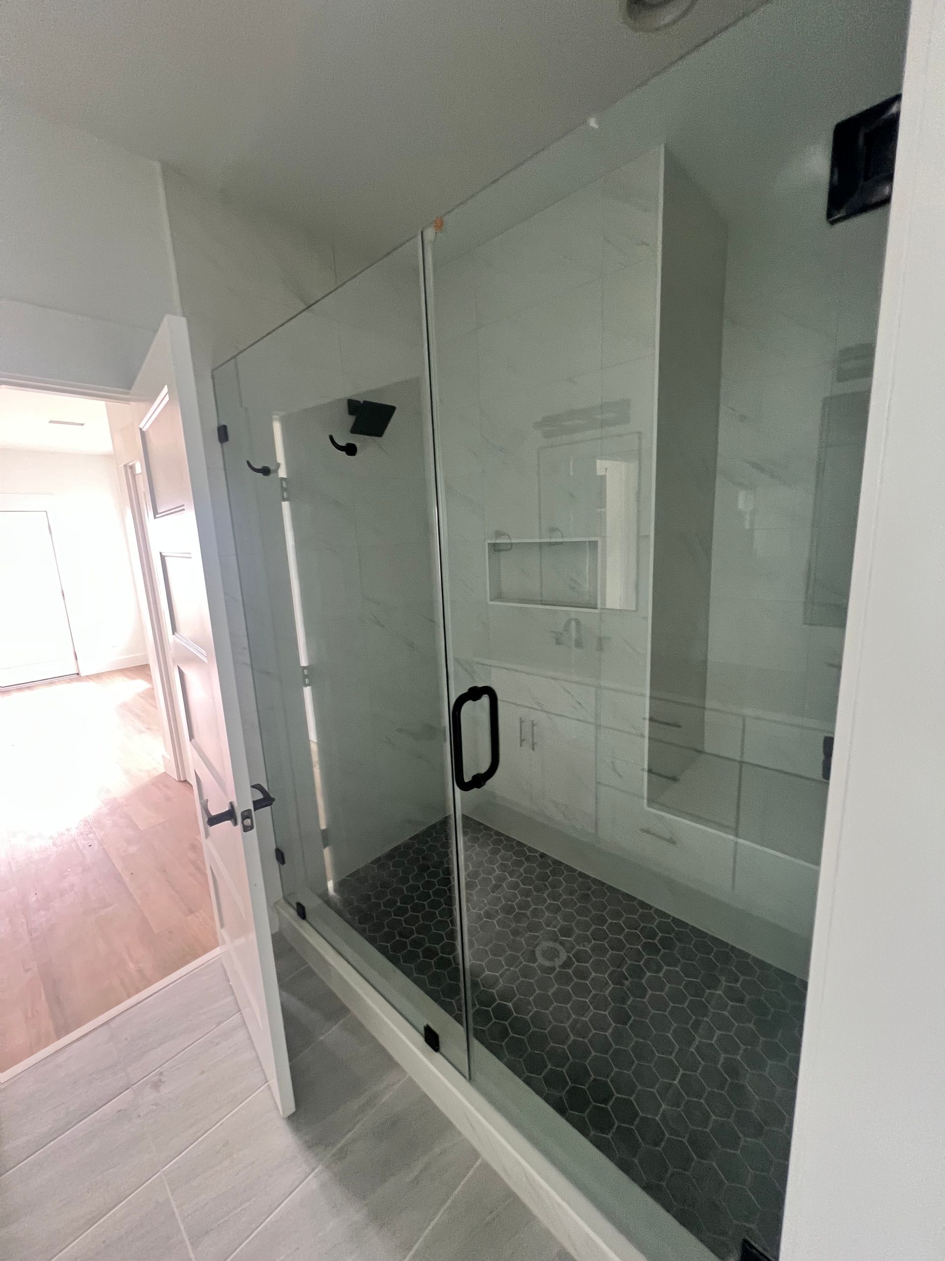 Glass shower with black hardware and dark tile floor. White walls and light wood floor visible.