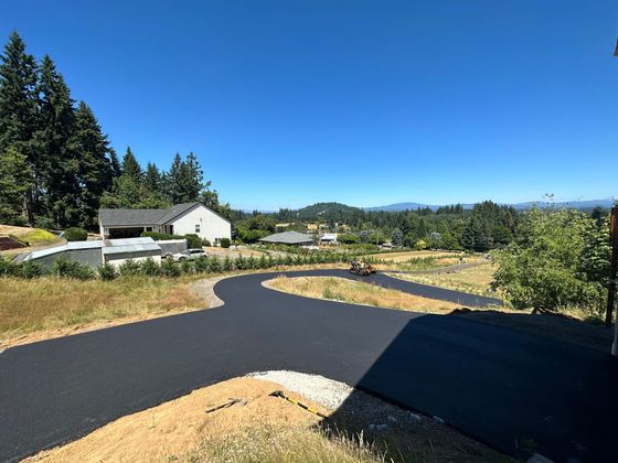 Curving paved road through a sunny rural landscape with trees, fields, and distant hills under a blue sky
