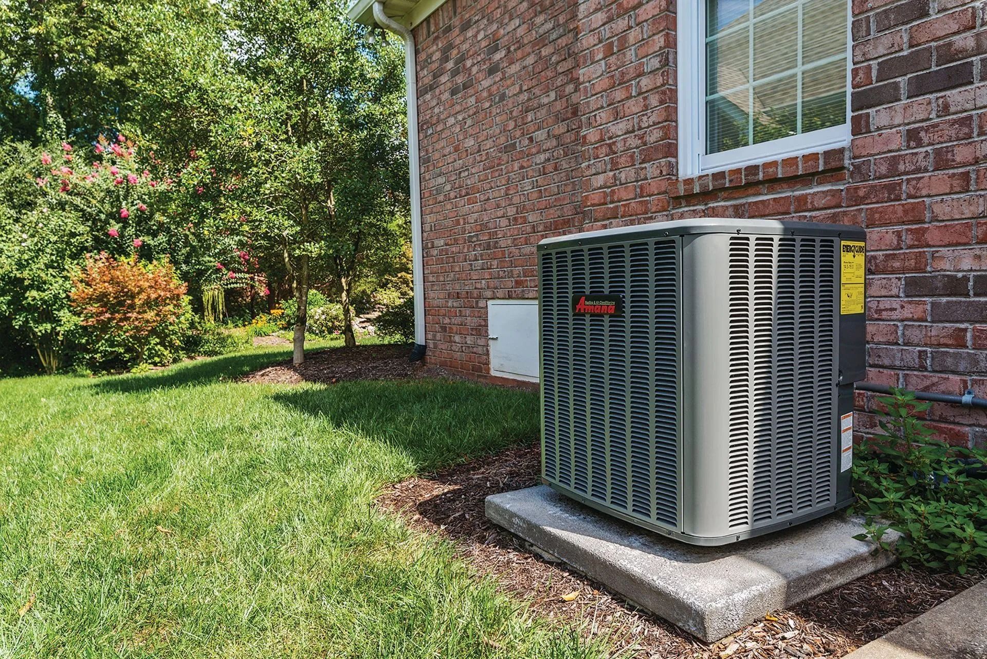 Air conditioning unit next to a brick house on a concrete pad, surrounded by grass and shrubs.