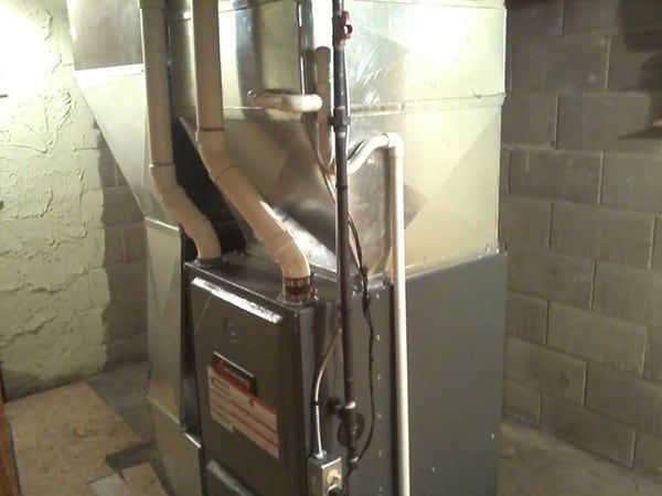 A furnace in a basement with metal ductwork and piping against a gray brick wall.
