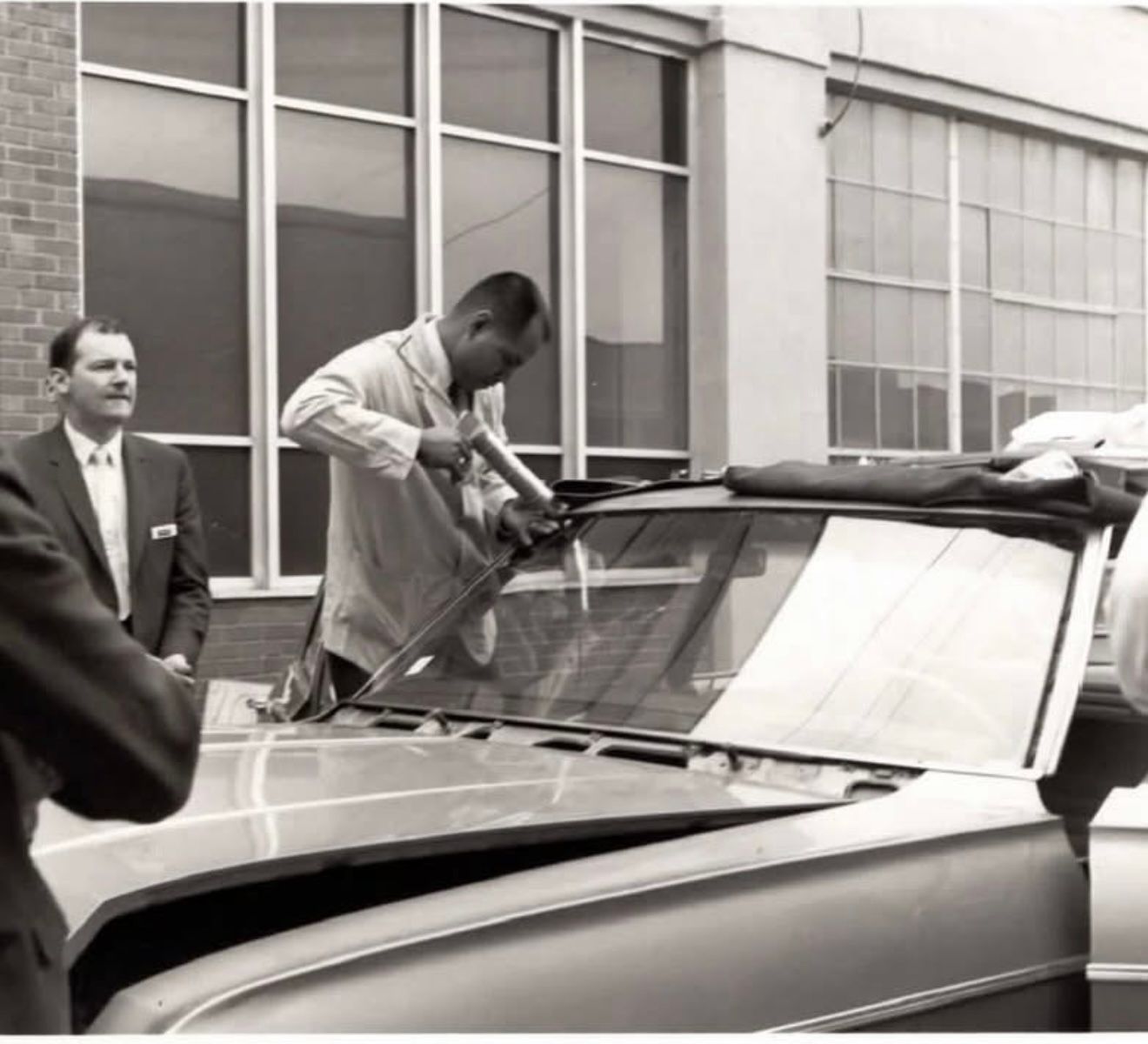 Man applying sealant to a car windshield outside a building. Two men watch nearby.